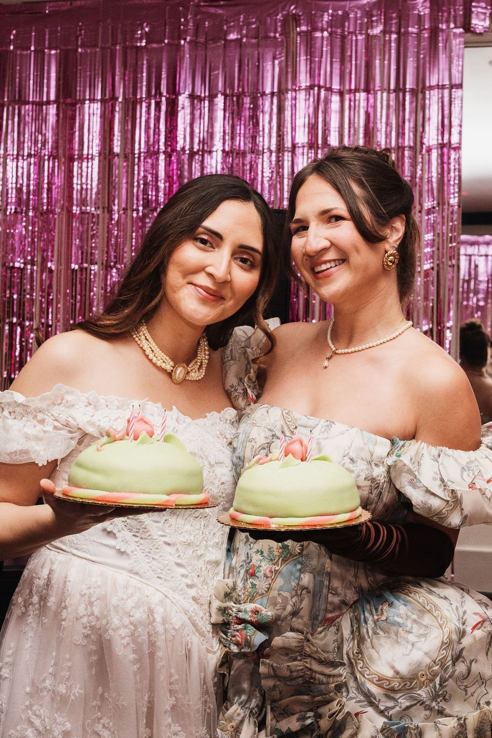 Two women in off-shoulder dresses holding green dome cakes at a pink tinsel backdrop celebration.
