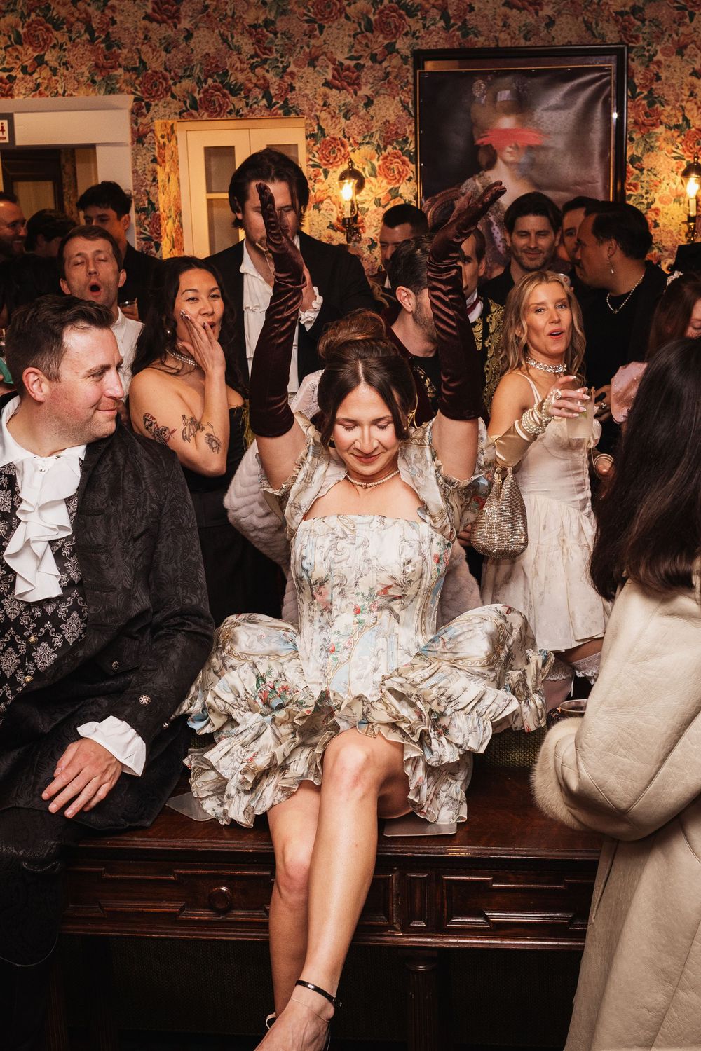 Woman in floral dress sits on table, dancing at a lively vintage-themed party with guests celebrating around her.