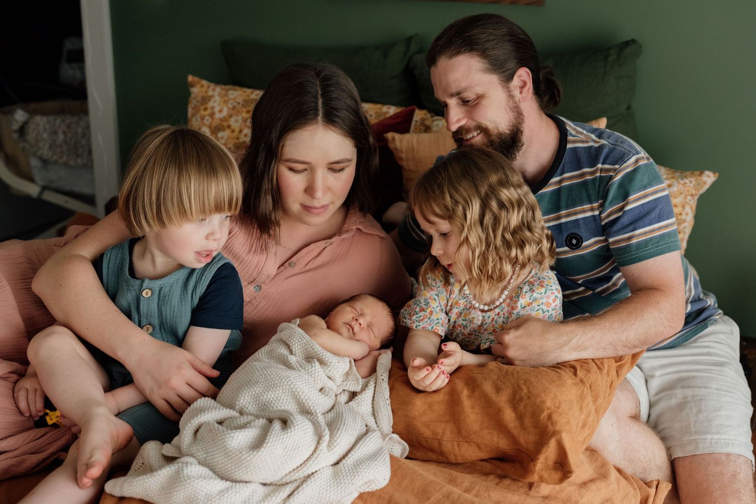 A couple smiles at their newborn baby on a bed with green walls and a New Zealand poster in a cozy bedroom.
