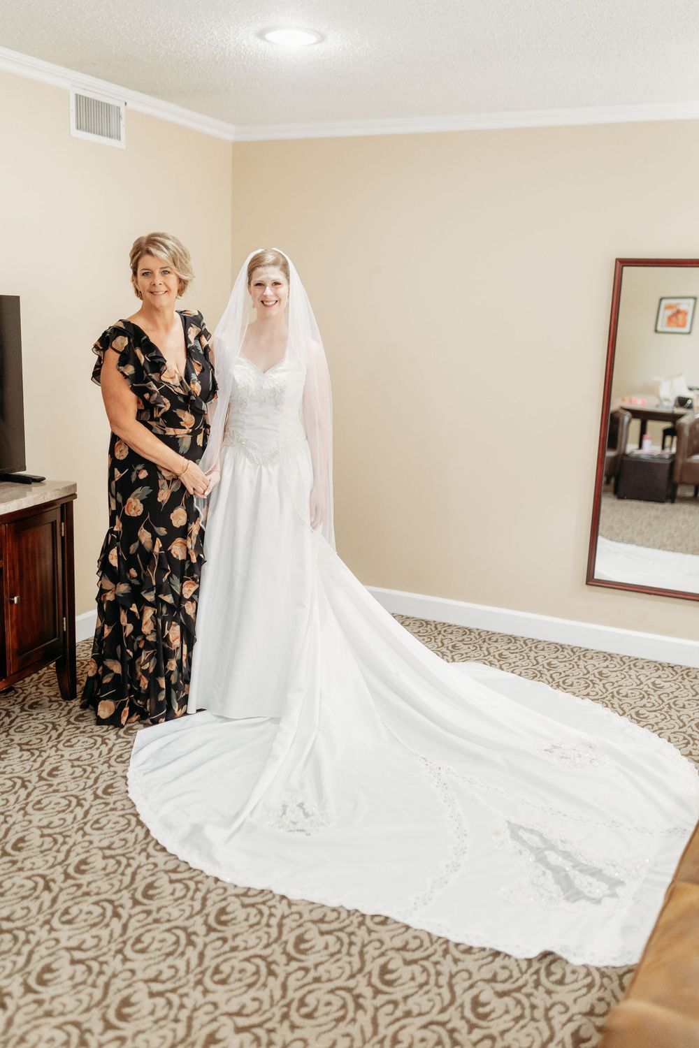 Bride in elegant white gown with long train stands with woman in floral dress in hotel bridal suite.