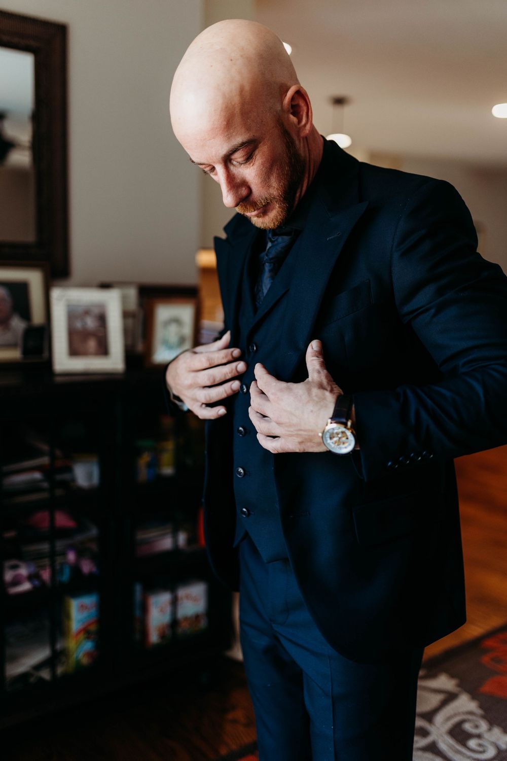 Bald groom in navy blue suit adjusting jacket lapels while getting ready for wedding ceremony.