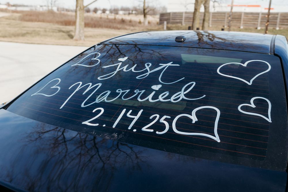 Just Married 2.14.25 written in white on dark car rear window with hearts.