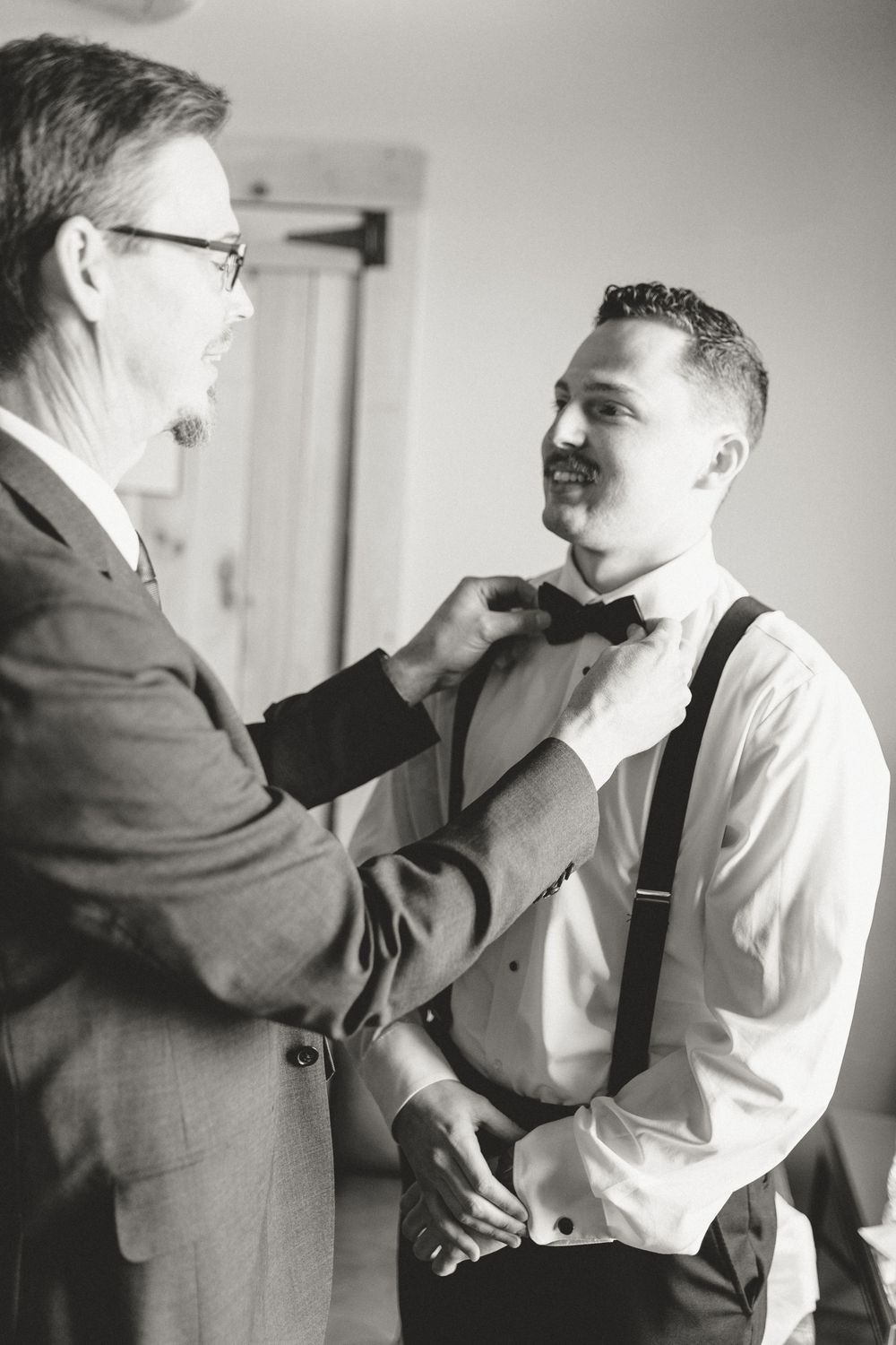 Black and white photo of older man adjusting groom's bow tie and suspenders before wedding.