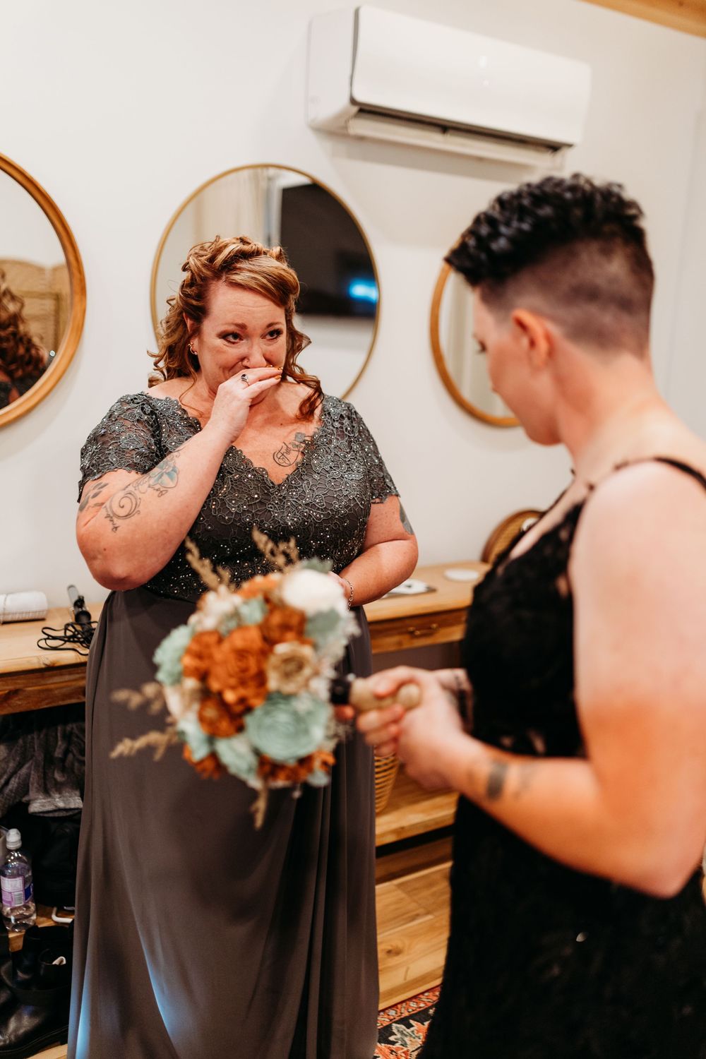 Emotional bridesmaid in gray gown holds bouquet while another person reads vows in rustic wedding prep room.