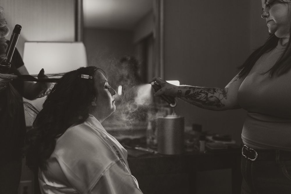 Black and white photo of a bride getting hair styled with steam visible in a dimly lit bridal preparation room.