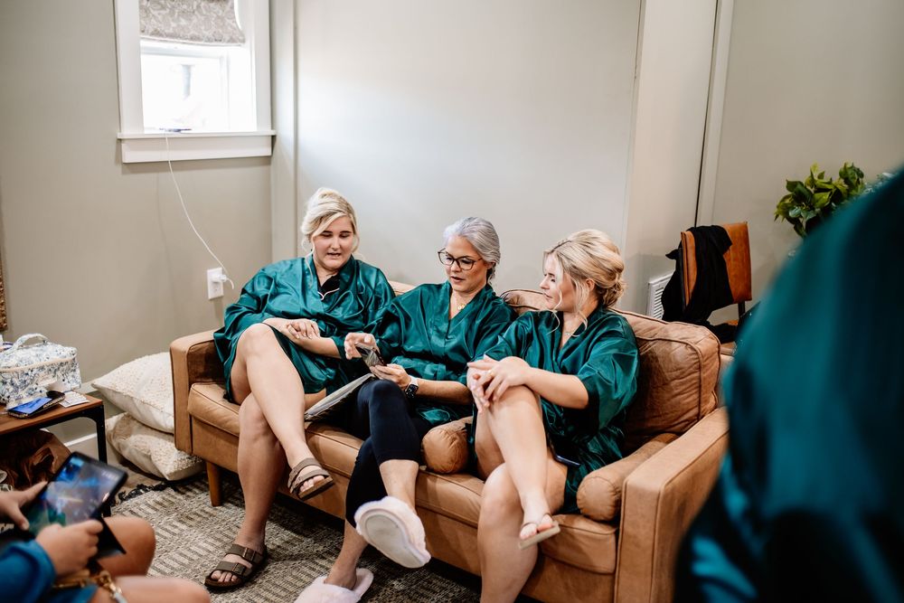 Three bridesmaids in teal robes laughing together on a couch while getting ready for a wedding.