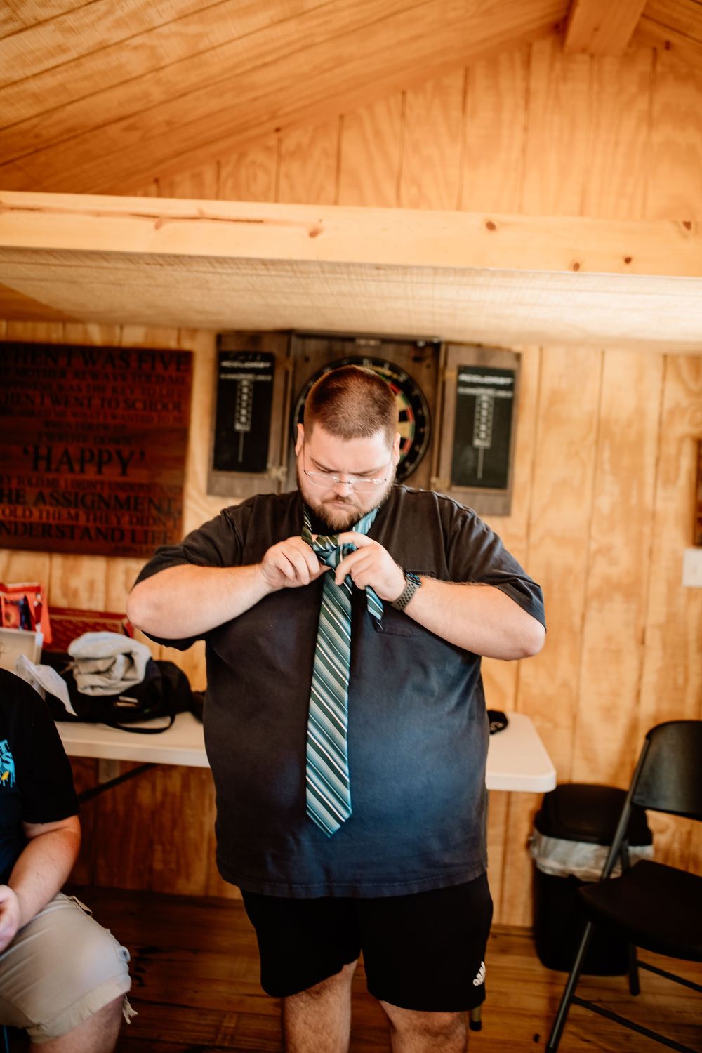 Man adjusting striped tie over dark shirt in rustic wooden room during wedding preparation.