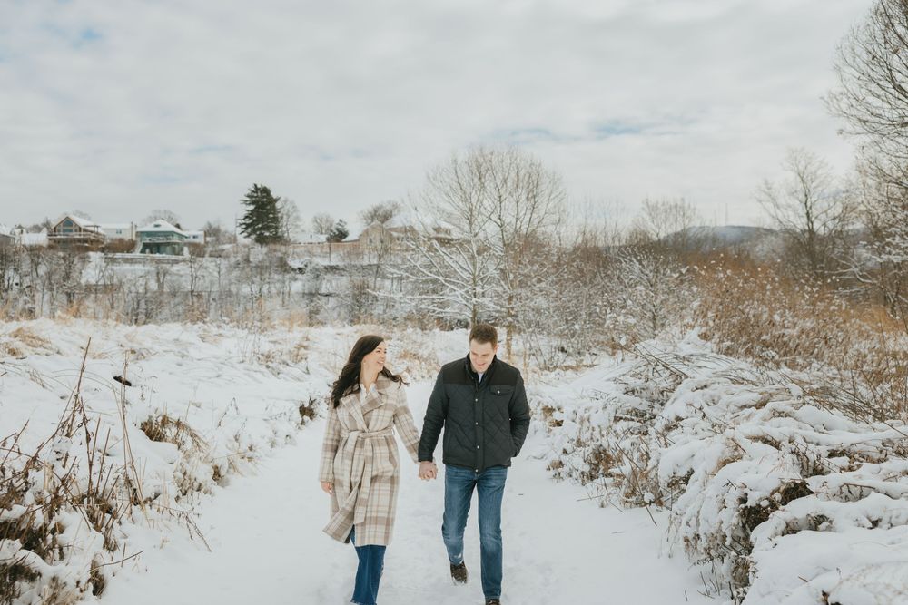 Couple walking hand in hand through a snowy winter landscape with bare trees and houses in the background.