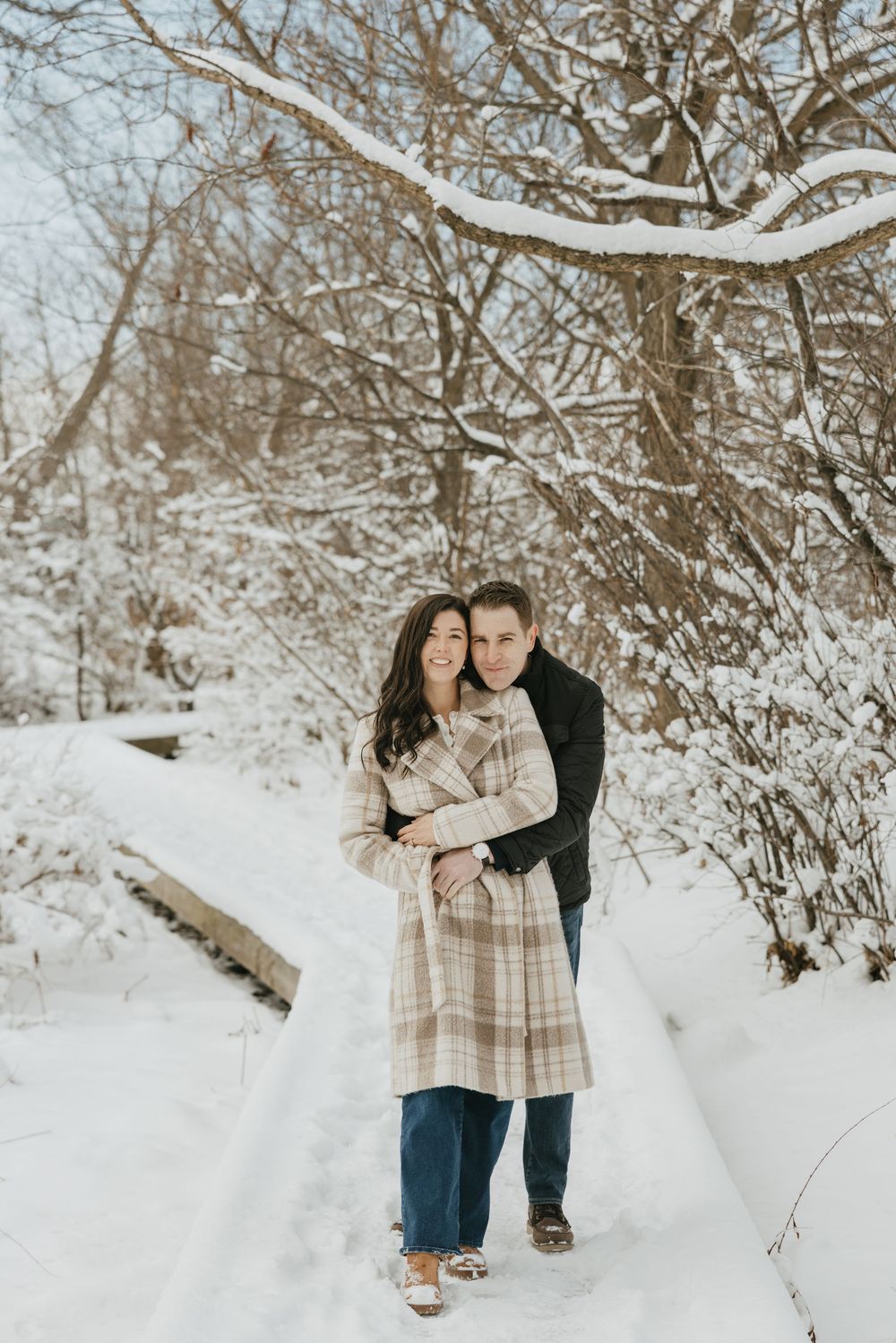 Couple embracing on a snowy winter path surrounded by snow-covered trees and branches.
