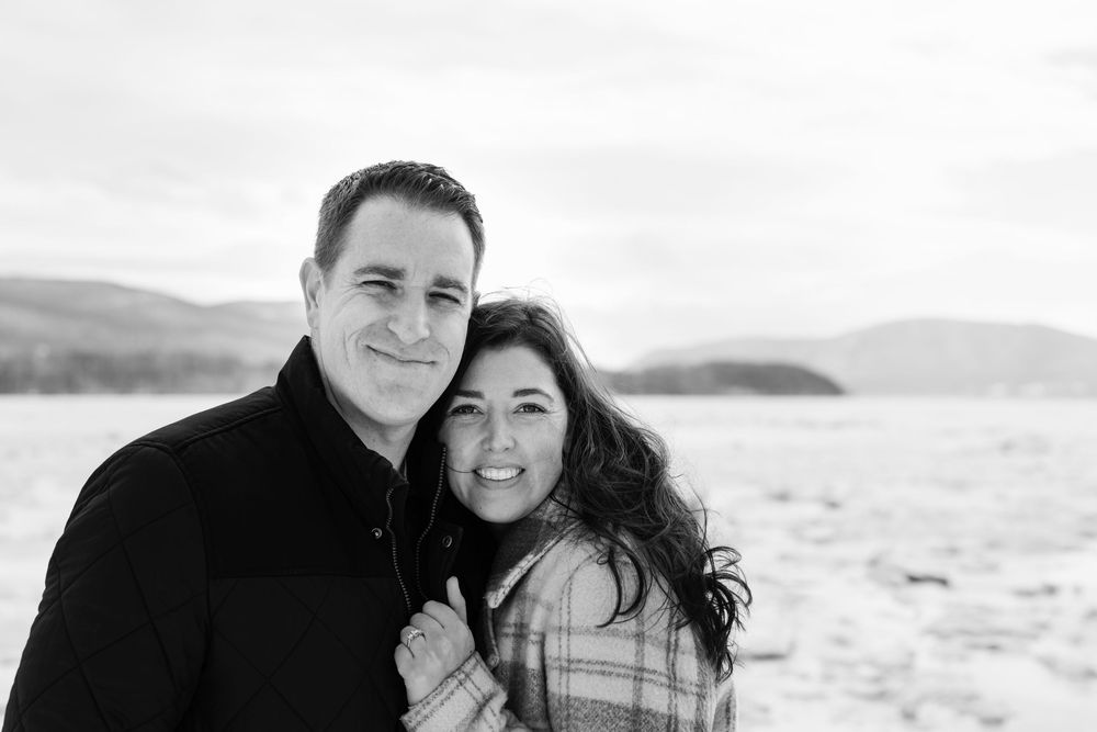 Black and white photo of a smiling couple embracing outdoors in winter with a snowy landscape and mountains behind them.