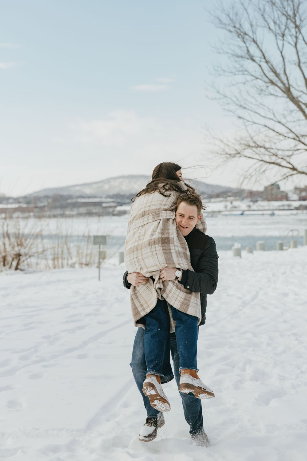 A man carries a woman wrapped in a plaid blanket through a snowy winter landscape near a frozen lake.