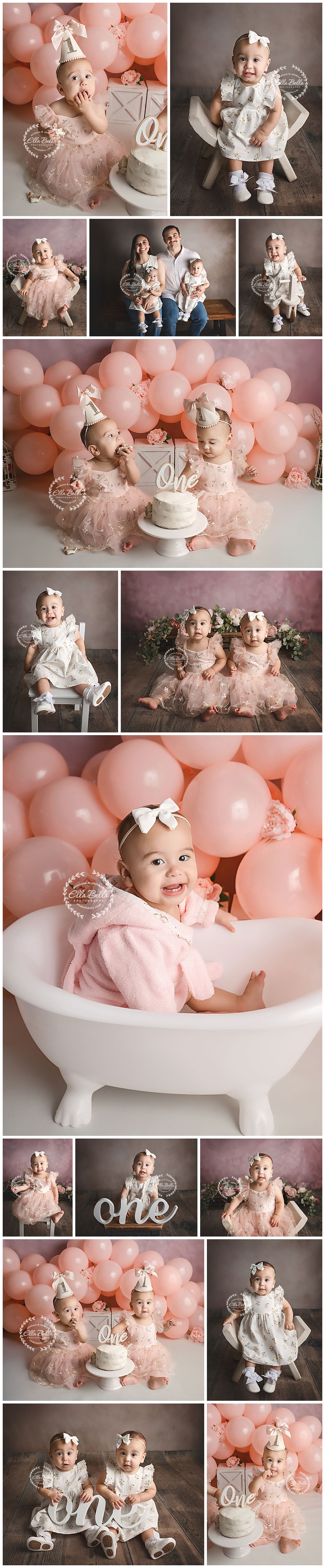 Twin baby girls in pink dresses celebrate first birthday with white cake and balloon backdrop.