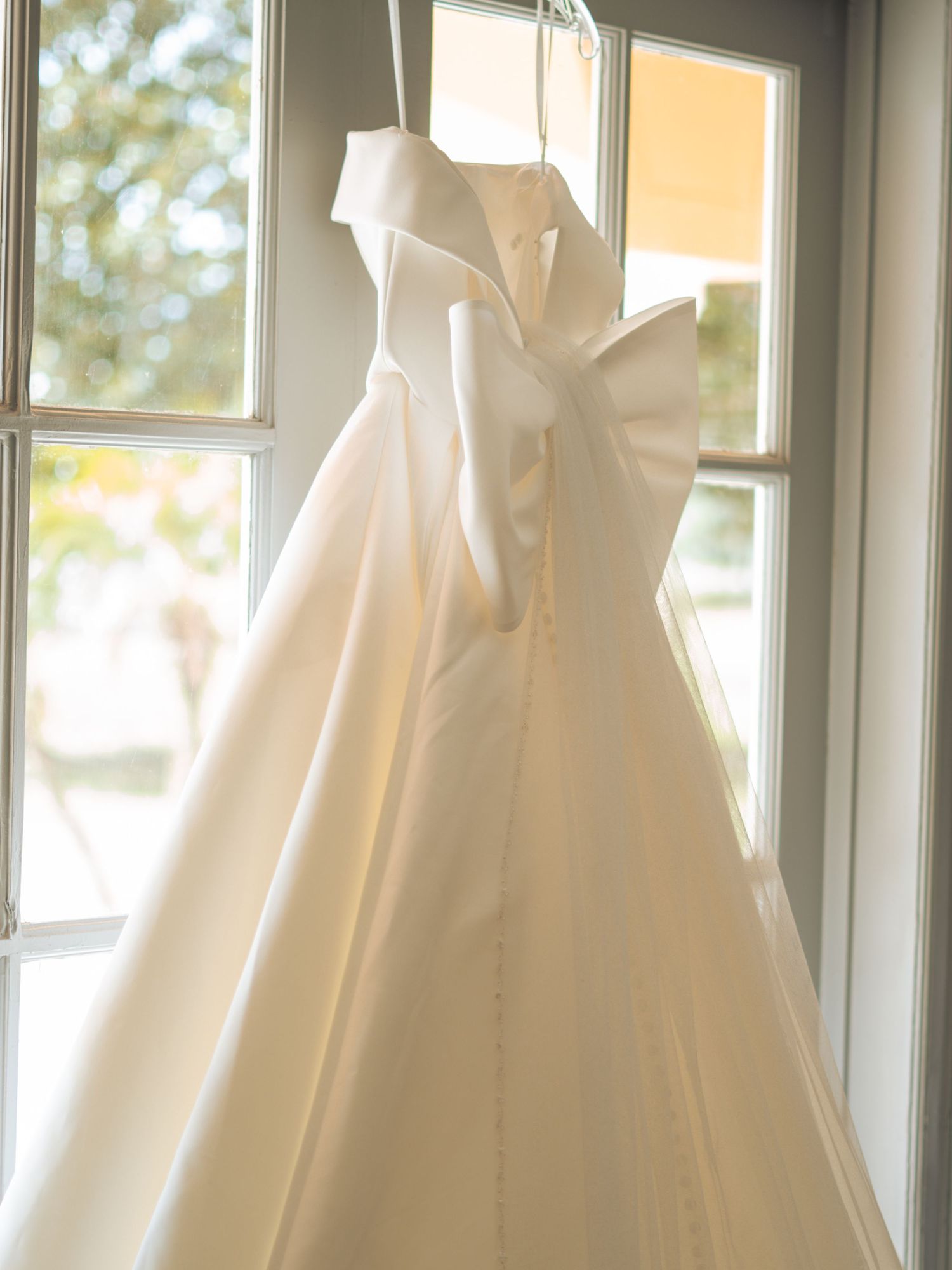 A white wedding dress hanging from an elegant chandelier in a beautifully decorated reception hall with gold table settings.