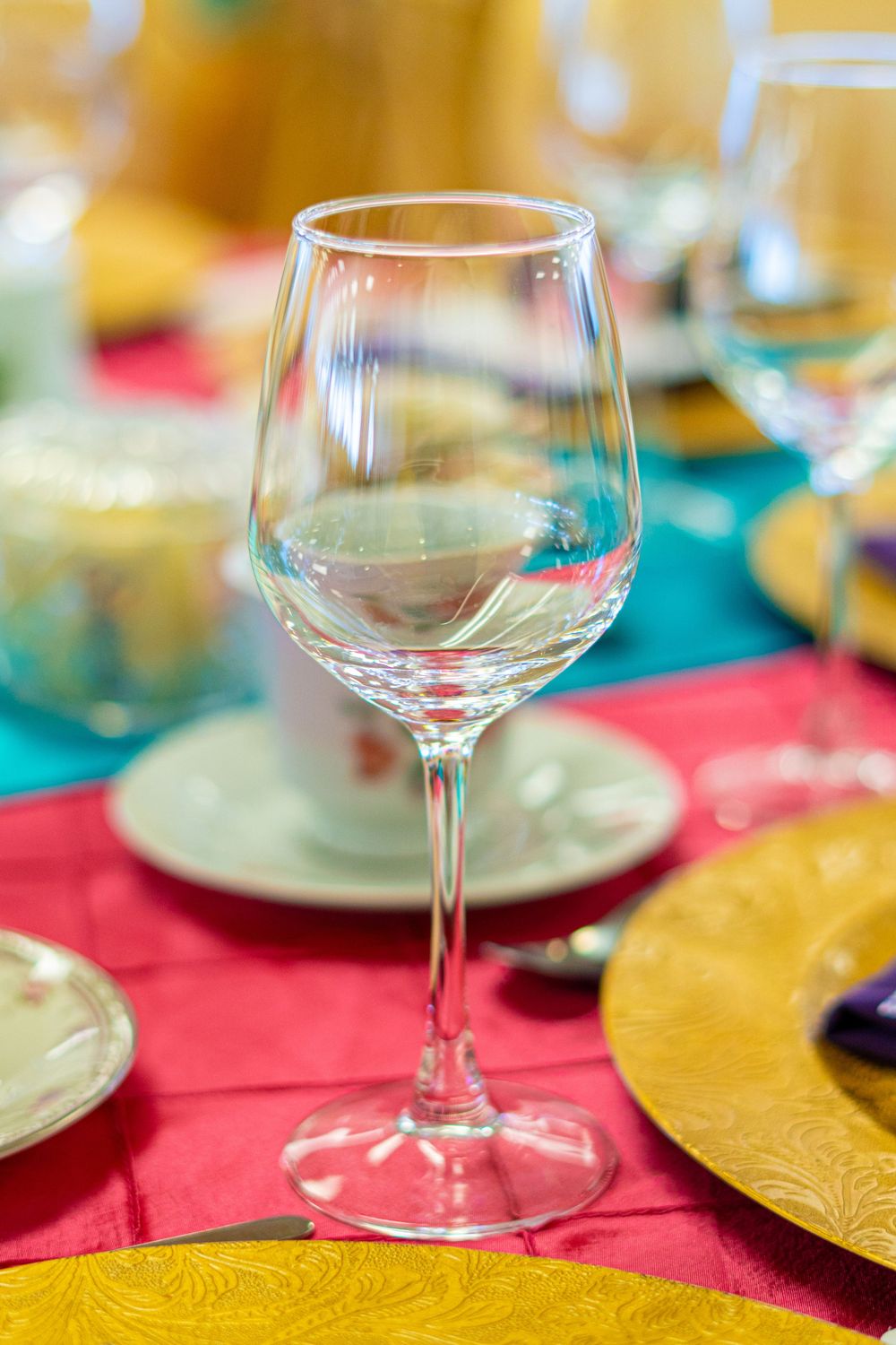 Empty wine glass on a colorful table setting with red and teal linens, gold charger plates, and blurred background.