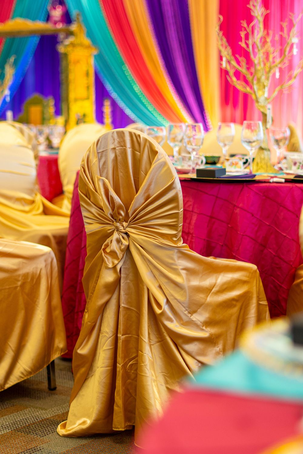 Gold satin chair covers with bow detail at a vibrant wedding reception with colorful draping.