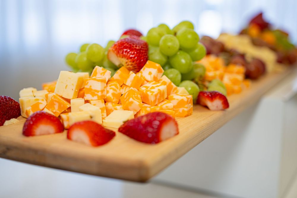 A wooden charcuterie board with cubed orange cheese, fresh strawberries, green grapes, and assorted fruits at a catered event.