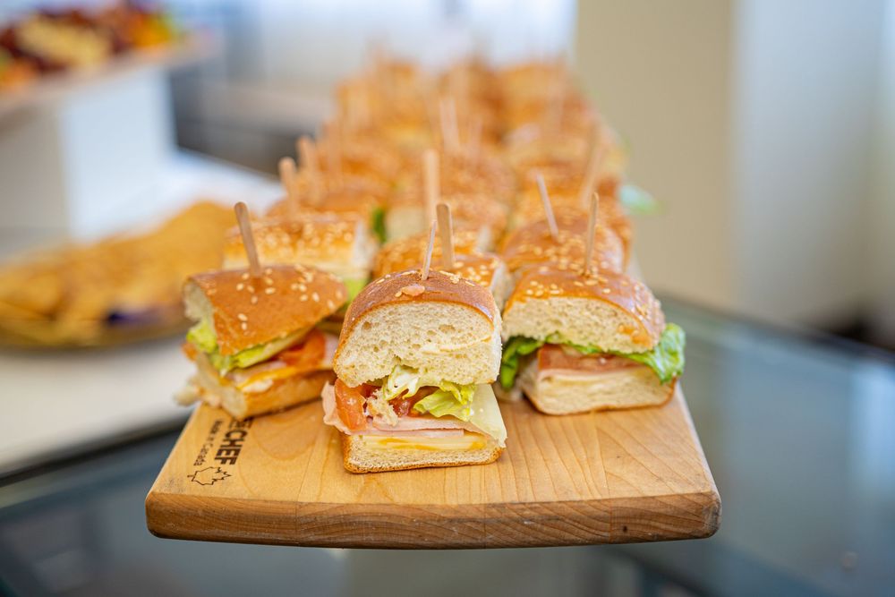 Rows of fresh deli sandwiches with lettuce and meat on sesame buns displayed on a wooden serving board.