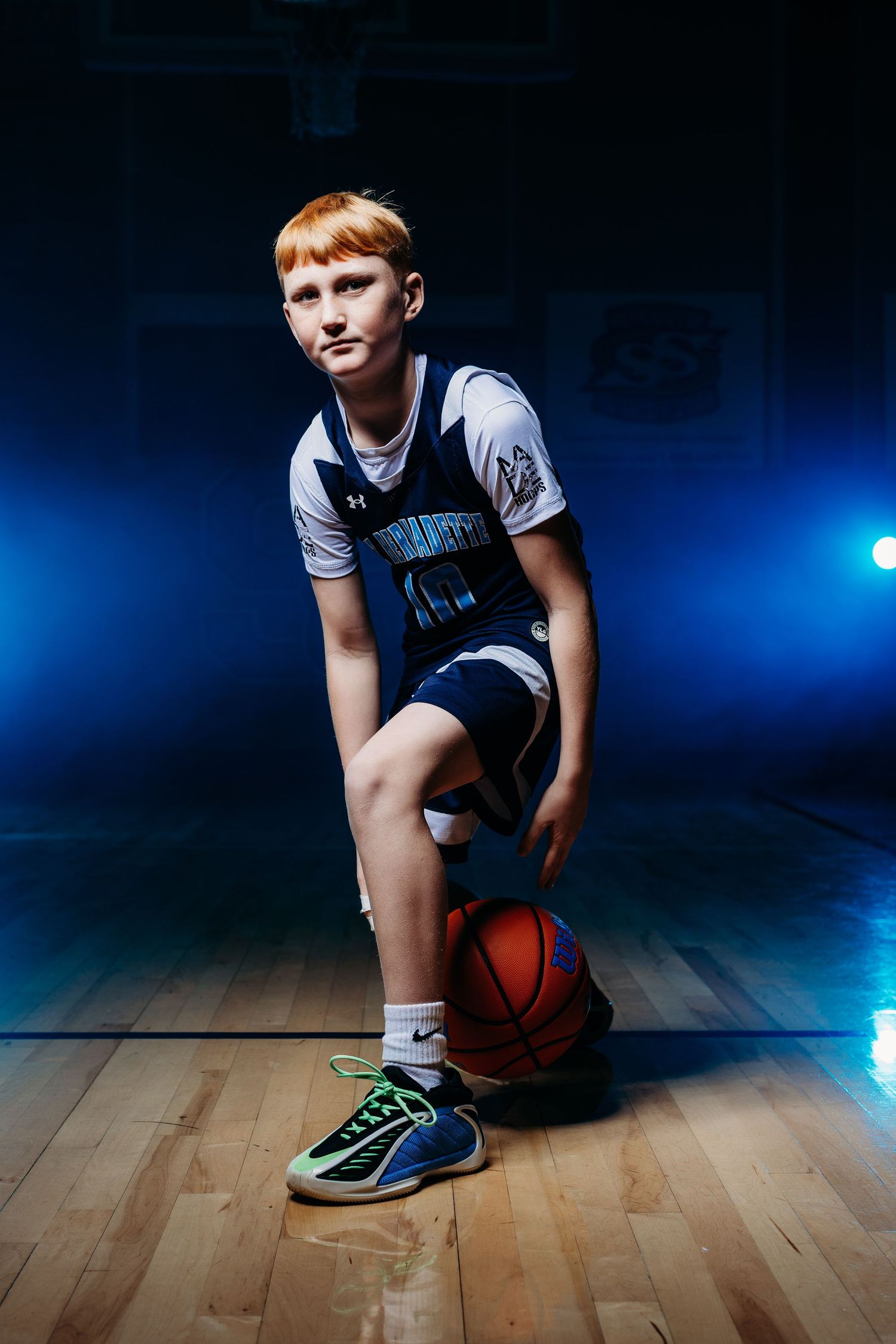 Young redhead boy in basketball uniform poses with ball on hardwood court under dramatic blue lighting.