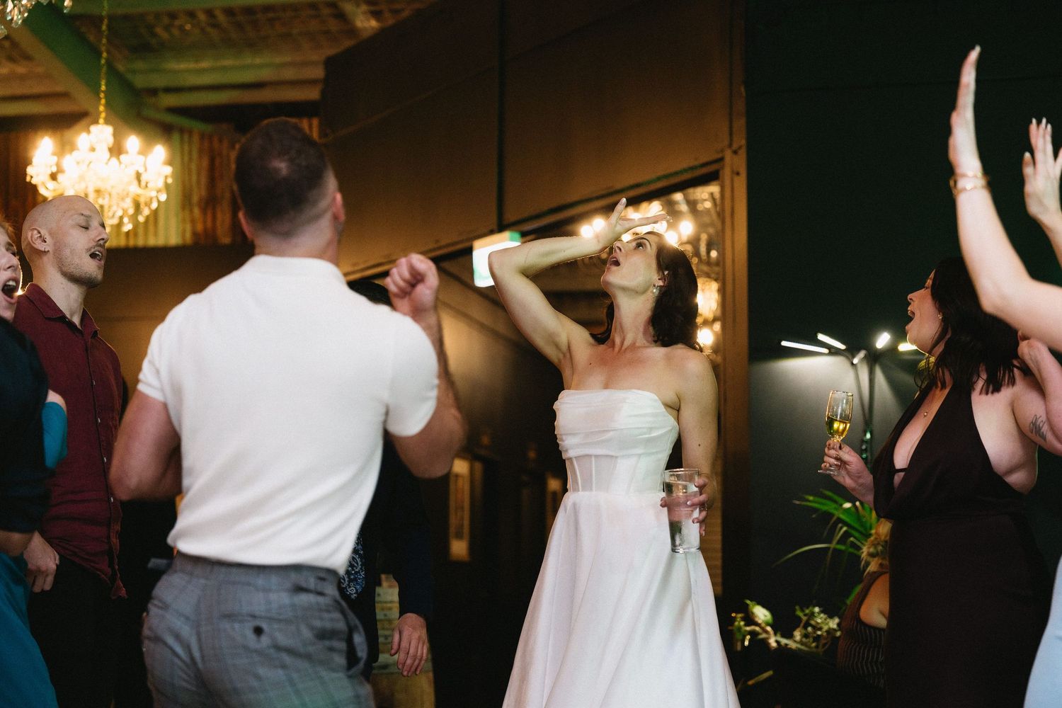Bride in white strapless gown dancing joyfully with guests at elegant wedding reception under chandelier lighting.