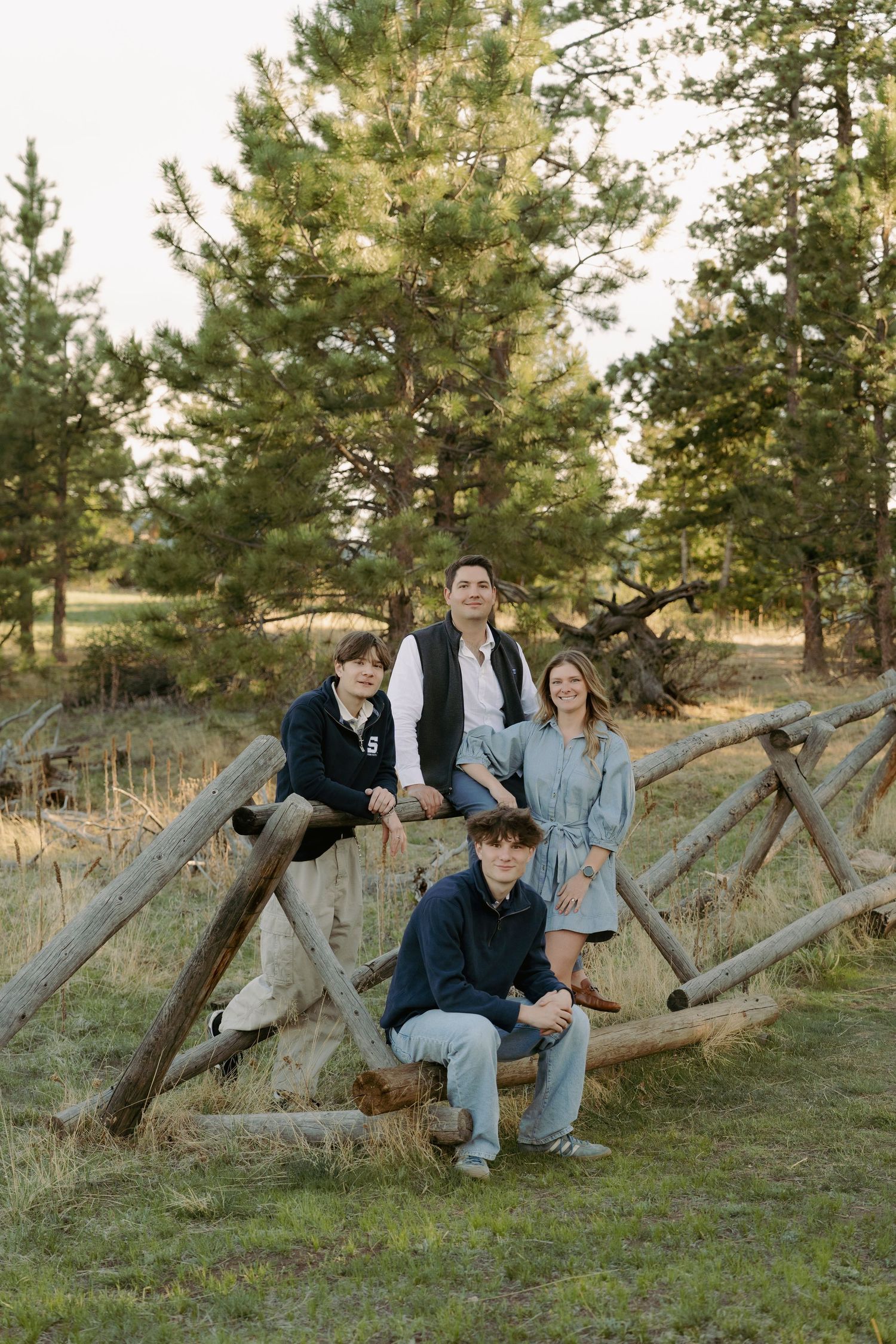 Family of four posing on rustic wooden fence surrounded by pine trees in an outdoor nature setting.