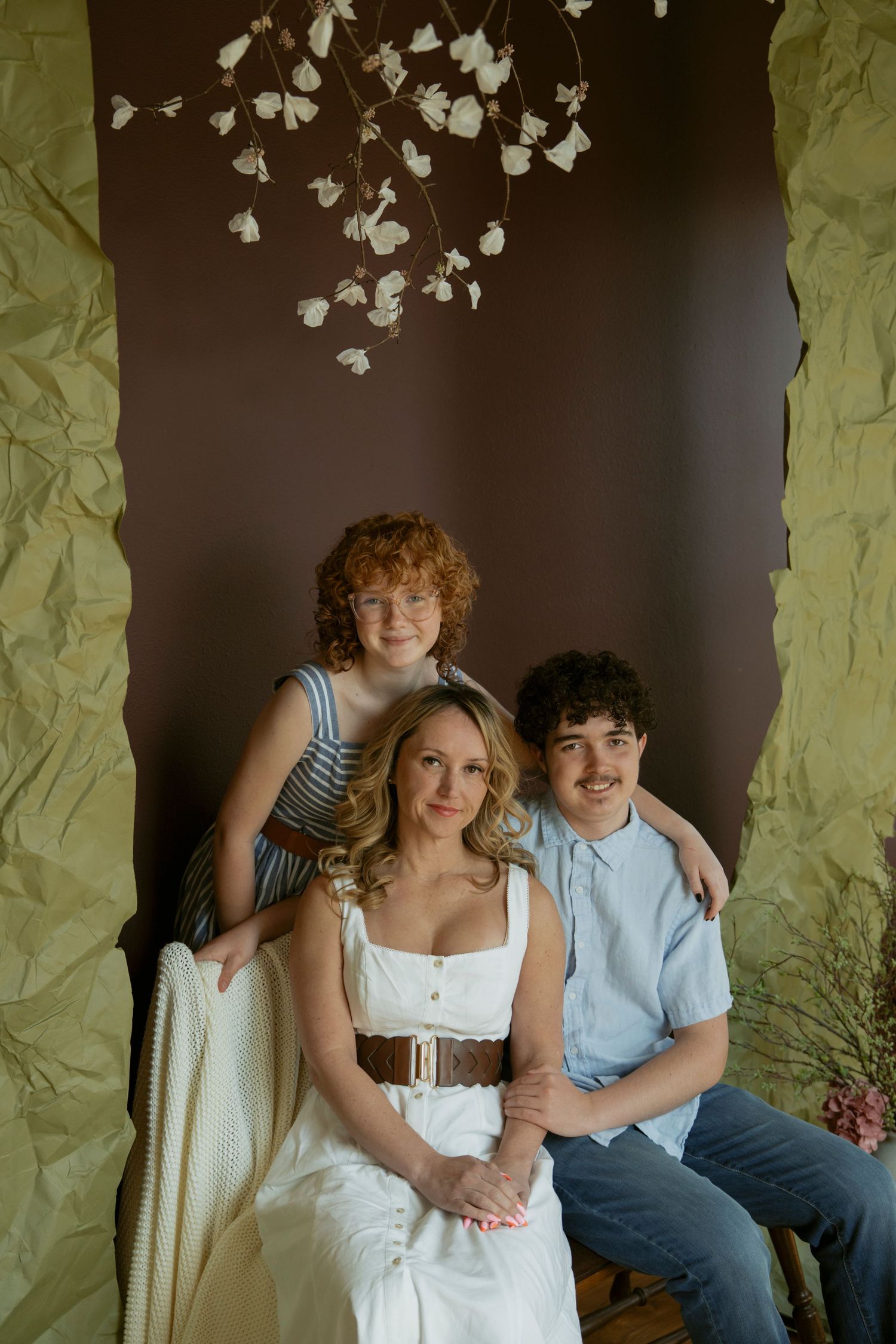A redhead girl in striped dress poses with a blonde woman in white, standing before a dark wall with white flowers.