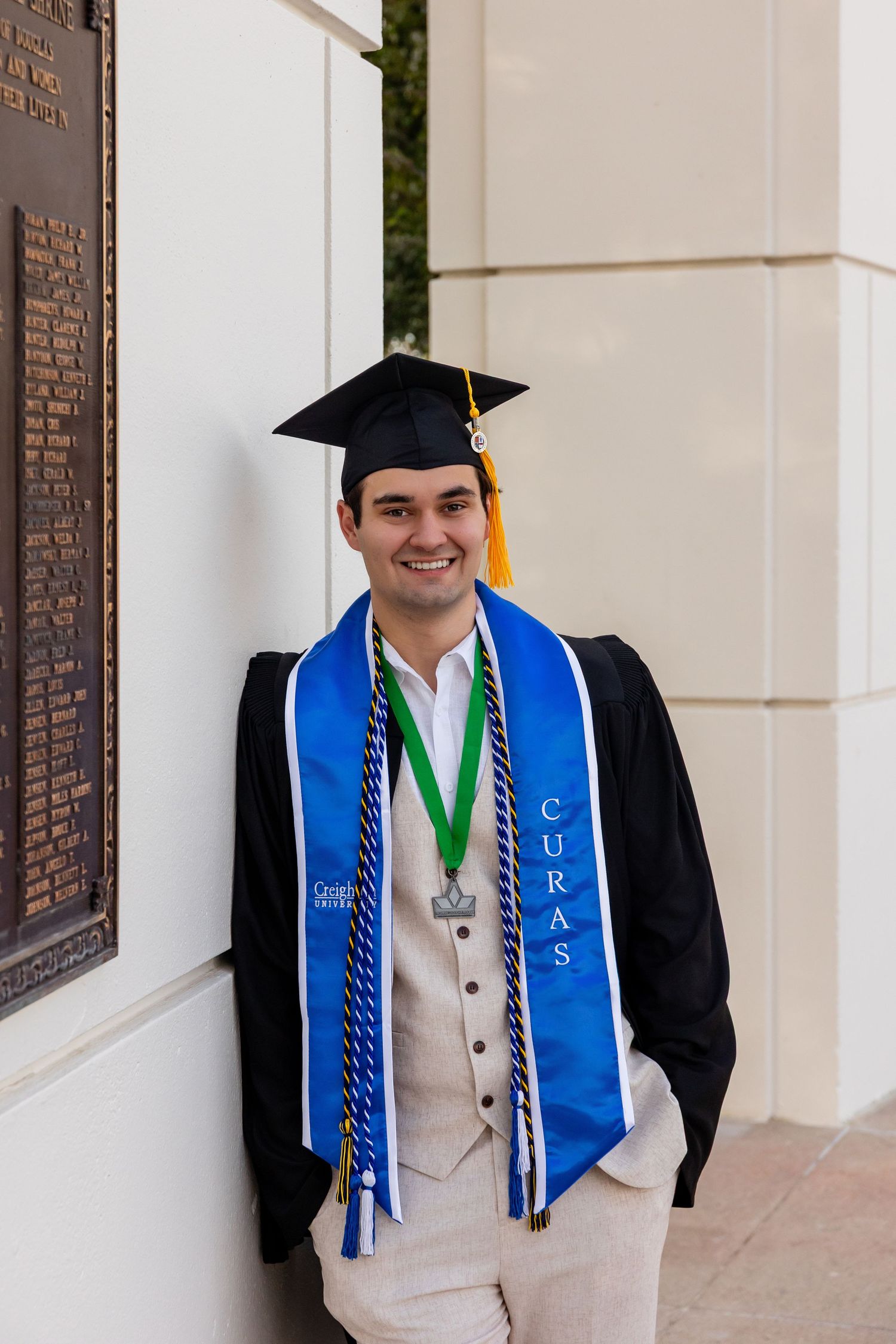 Smiling male Creighton graduate in black cap and gown with blue CURAS stole and medal posing outdoors at Memorial Park in Omaha, NE.