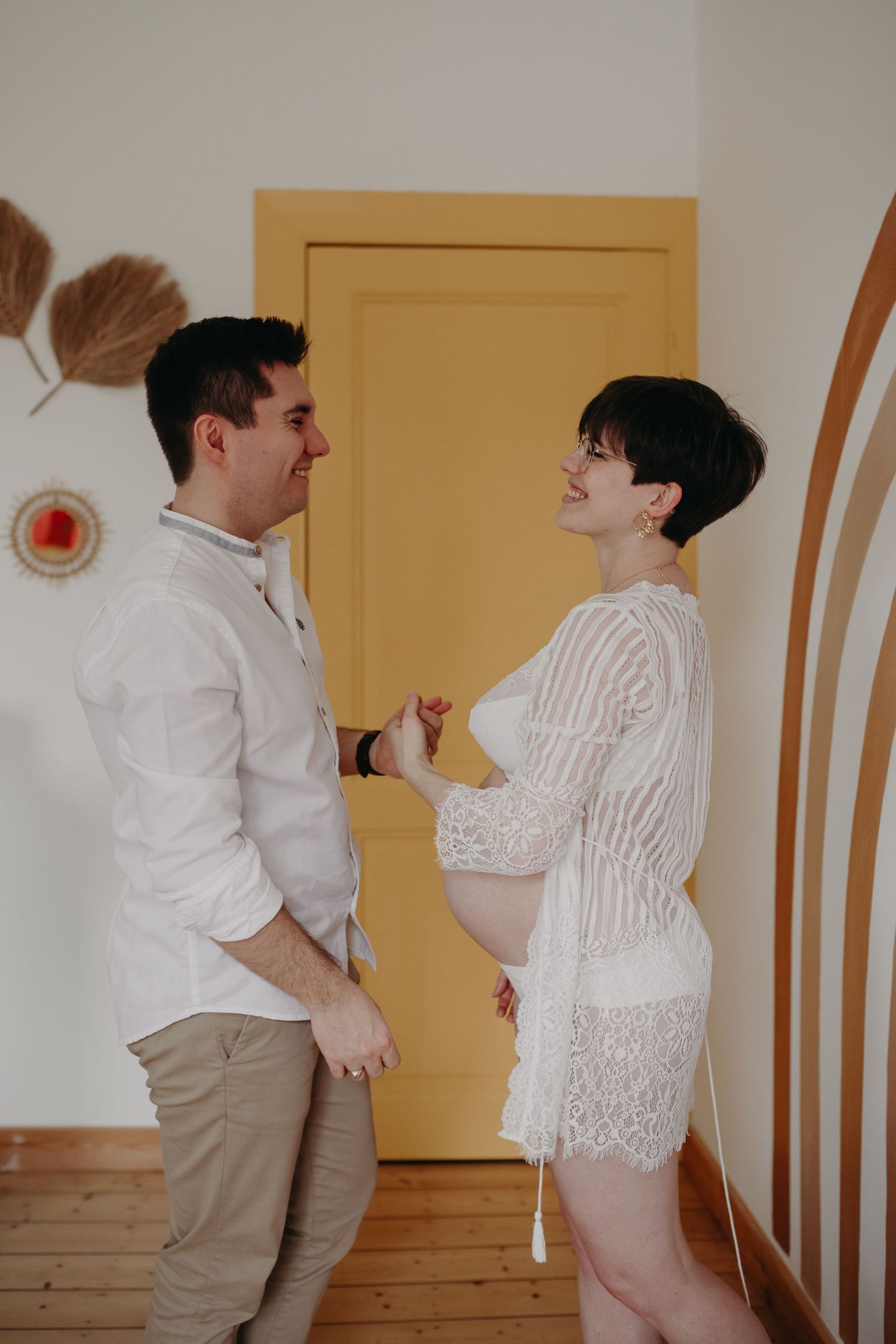 Smiling pregnant couple holding hands in front of a yellow door in a bright room.