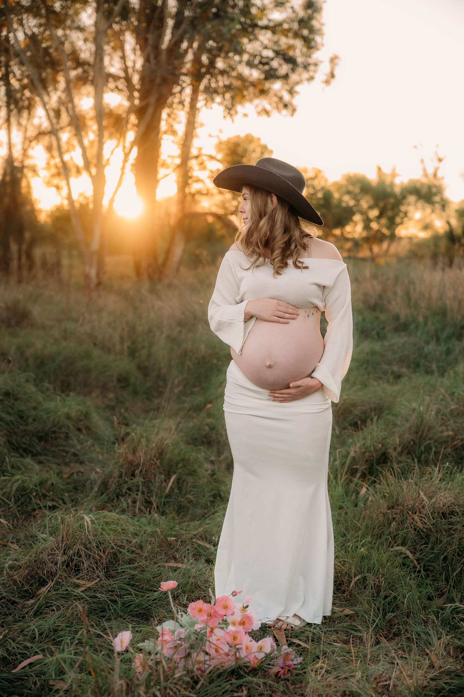 Pregnant woman in white dress and cowboy hat cradling baby bump at golden sunset in a green field.