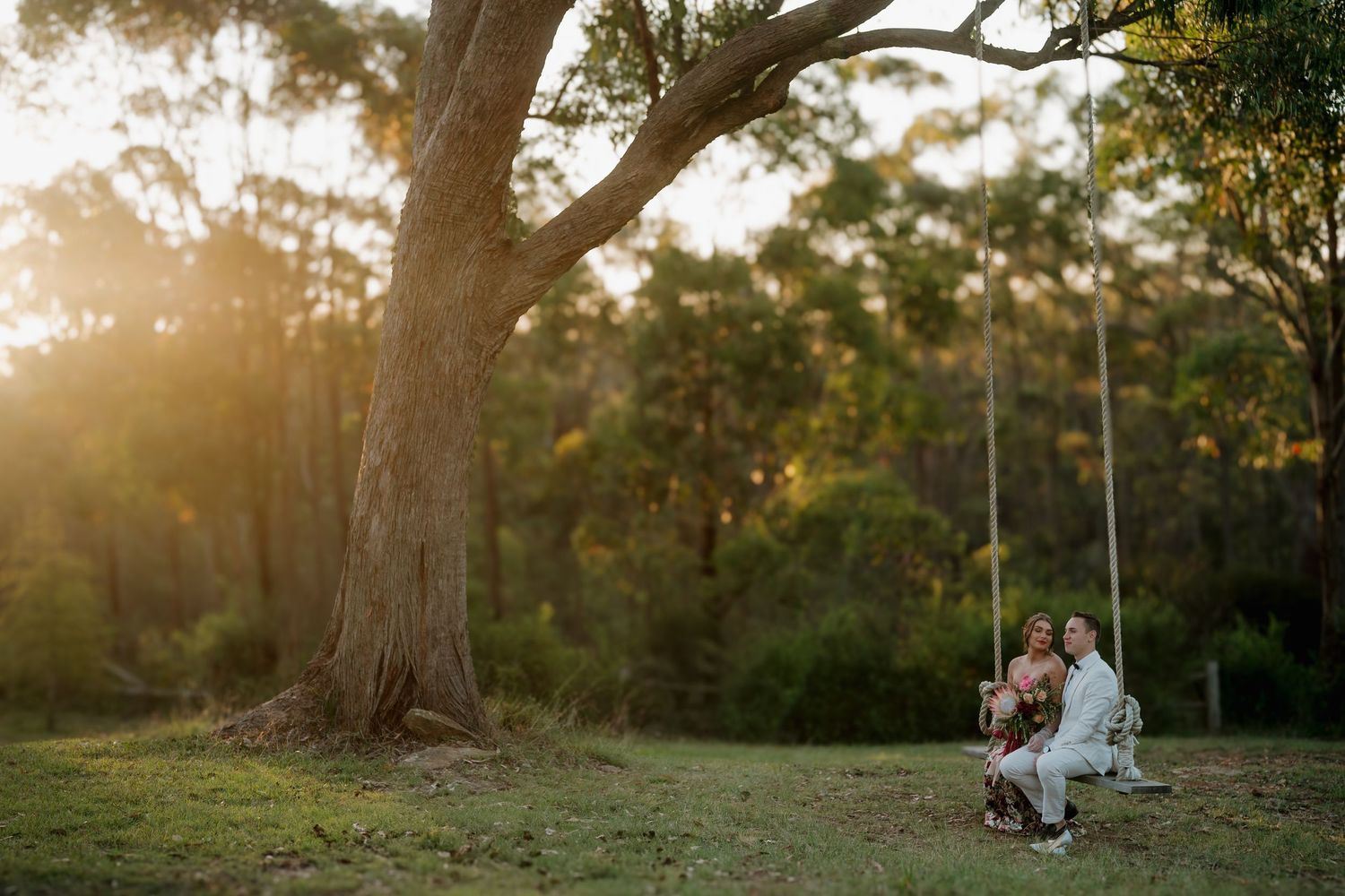 Couple sitting on a rope swing under a large tree at golden hour in a lush Australian bush setting.