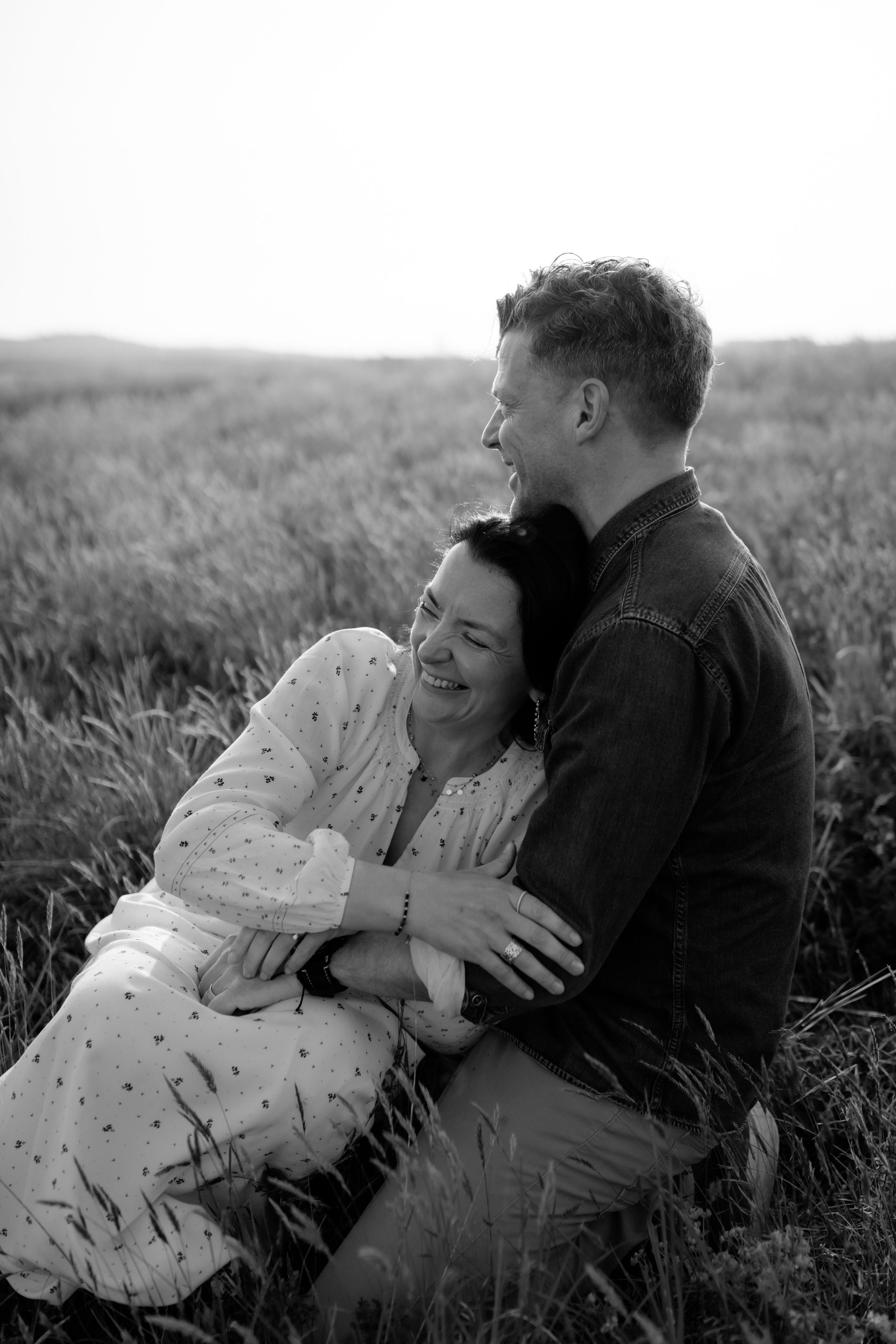 Black and white photo of a laughing couple embracing in a field, sharing a joyful romantic moment outdoors.