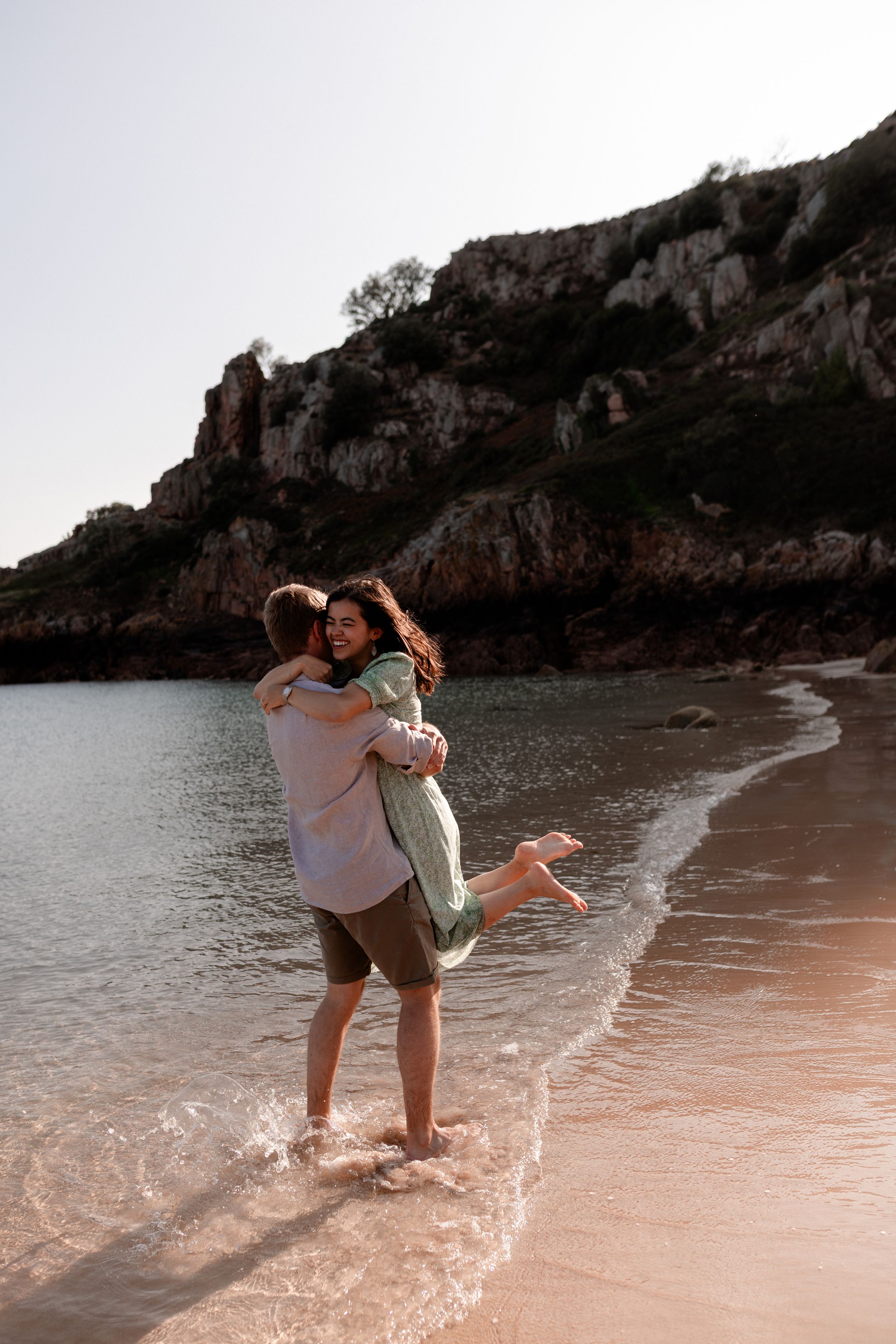Couple embracing on a sandy beach with rocky cliffs and ocean waves in the background.