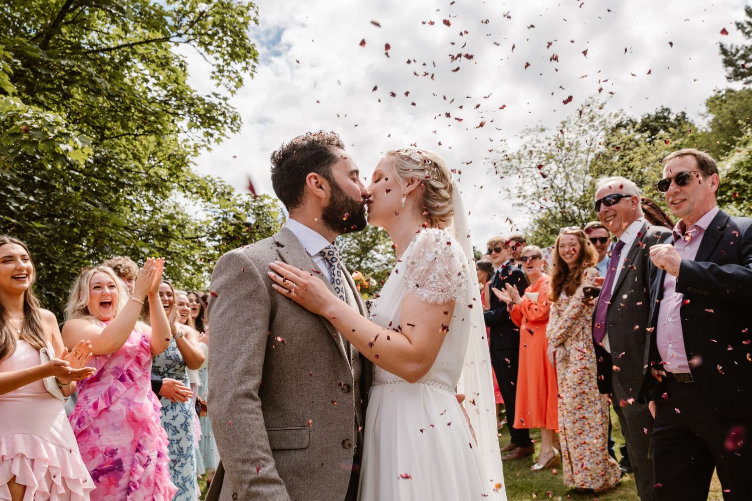 Bride and groom embrace in a lush garden, she laughs joyfully in a lace gown as he holds her in a blue suit.