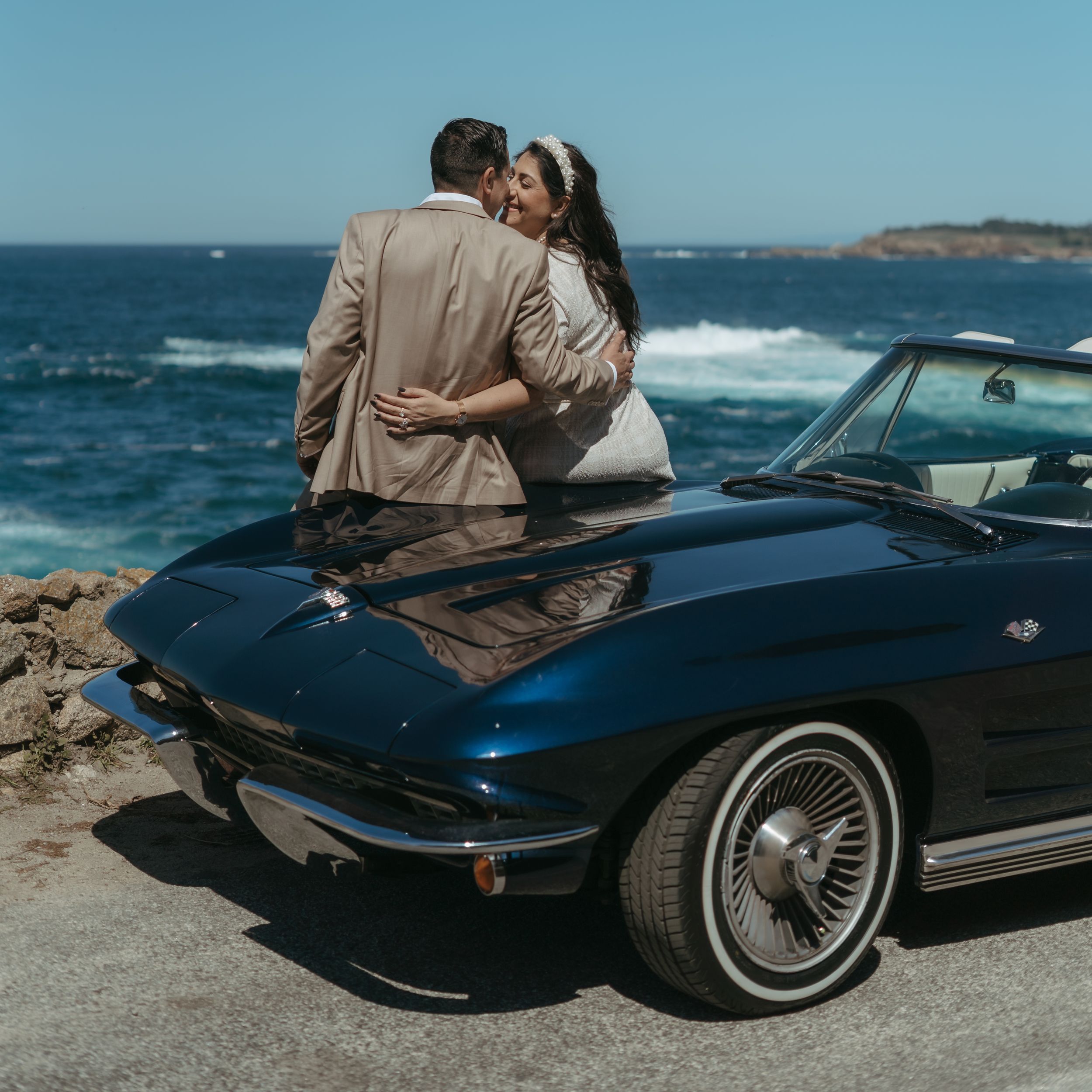 Couple holding hands beside a vintage blue convertible on a coastal cliff overlooking turquoise ocean waves.