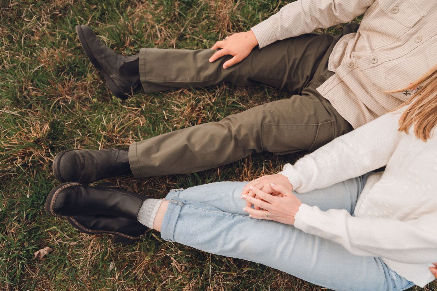 Couple sitting on grass holding hands, wearing dark boots, olive pants and light jeans, outdoor fall setting.