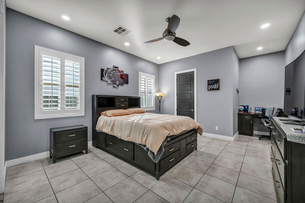 Bedroom with tile floors, ceiling fan, plantation shutters, dark wood furniture, and gray walls.