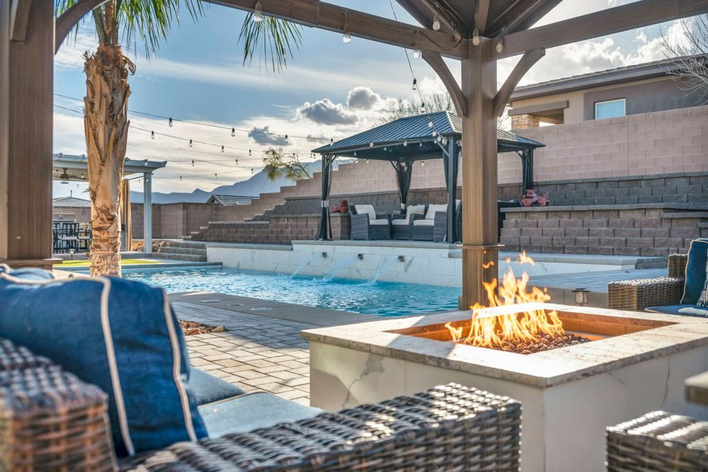 Outdoor patio with fire pit, wicker furniture, pergola, and sparkling pool at dusk.