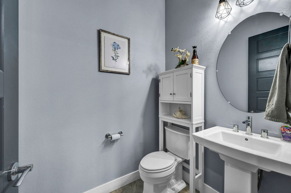 Clean powder room with gray walls, white pedestal sink, round mirror, and over-toilet storage cabinet.