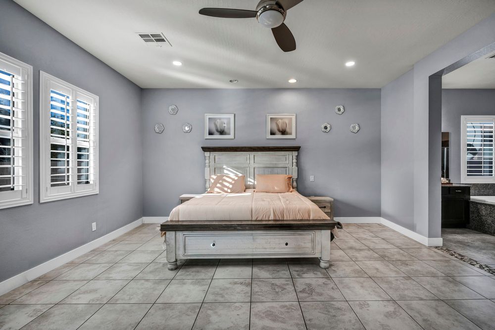 Spacious master bedroom with gray walls, tile floors, ceiling fan, and plantation shutters.
