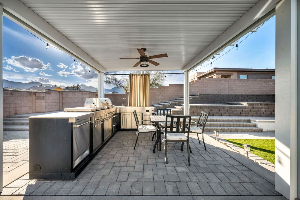 Covered outdoor kitchen with built-in grill, ceiling fan, patio dining set, and brick wall backdrop.