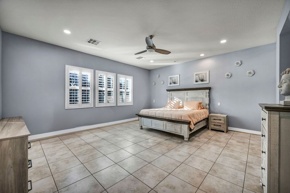 Large bedroom with gray walls, light tile floors, ceiling fan, plantation shutters, and wooden bed frame.