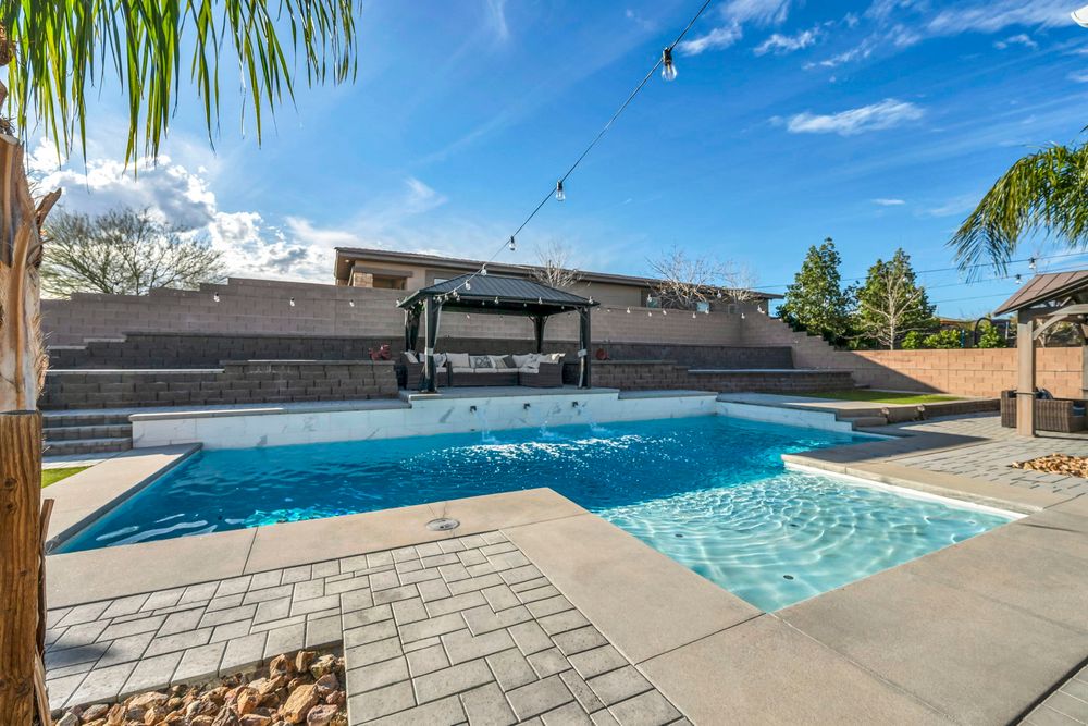 Backyard pool with blue water, paver surround, pergola seating area, and block wall privacy fence.