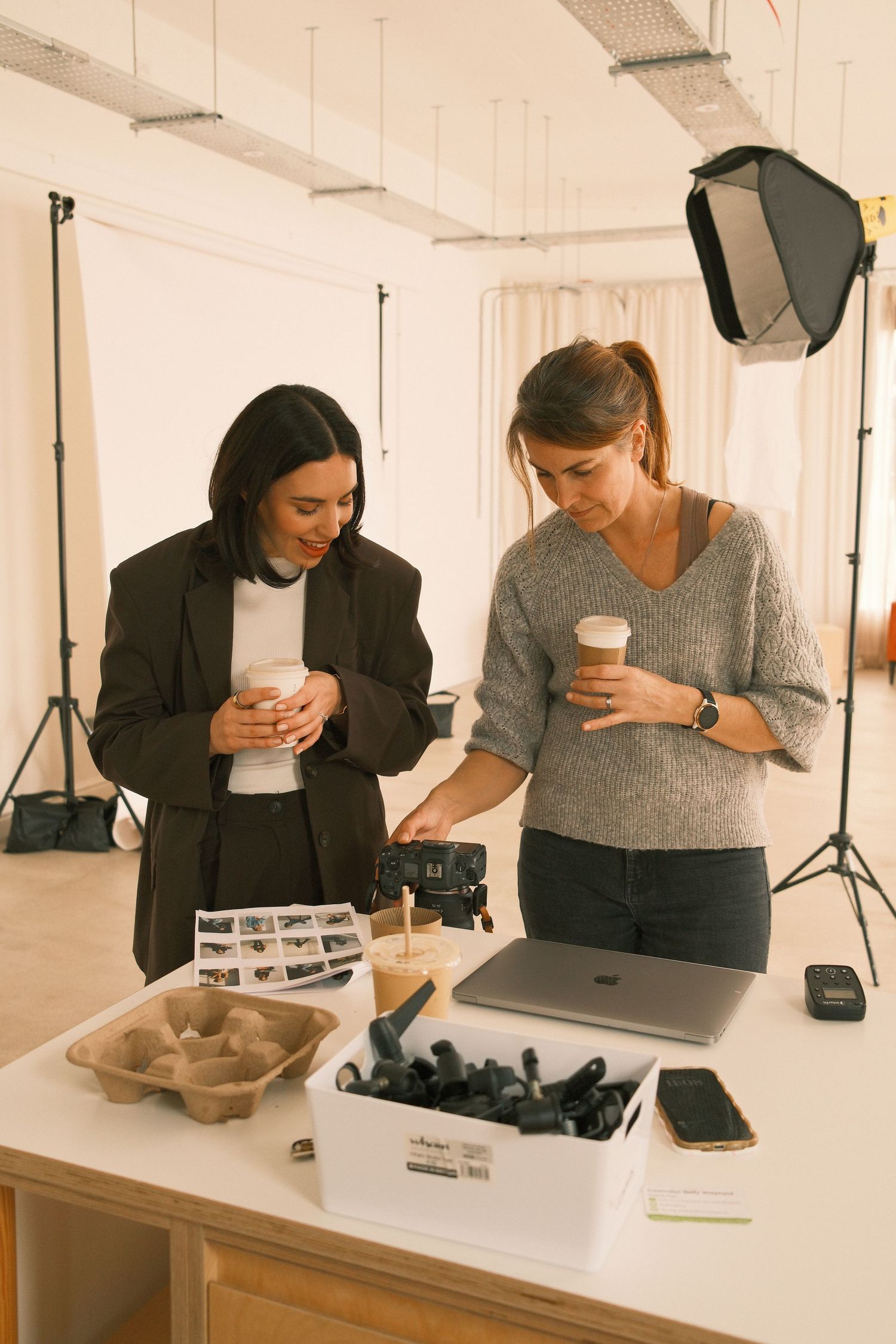 Two women holding coffee cups review photos and camera equipment on a table in a professional photography studio.