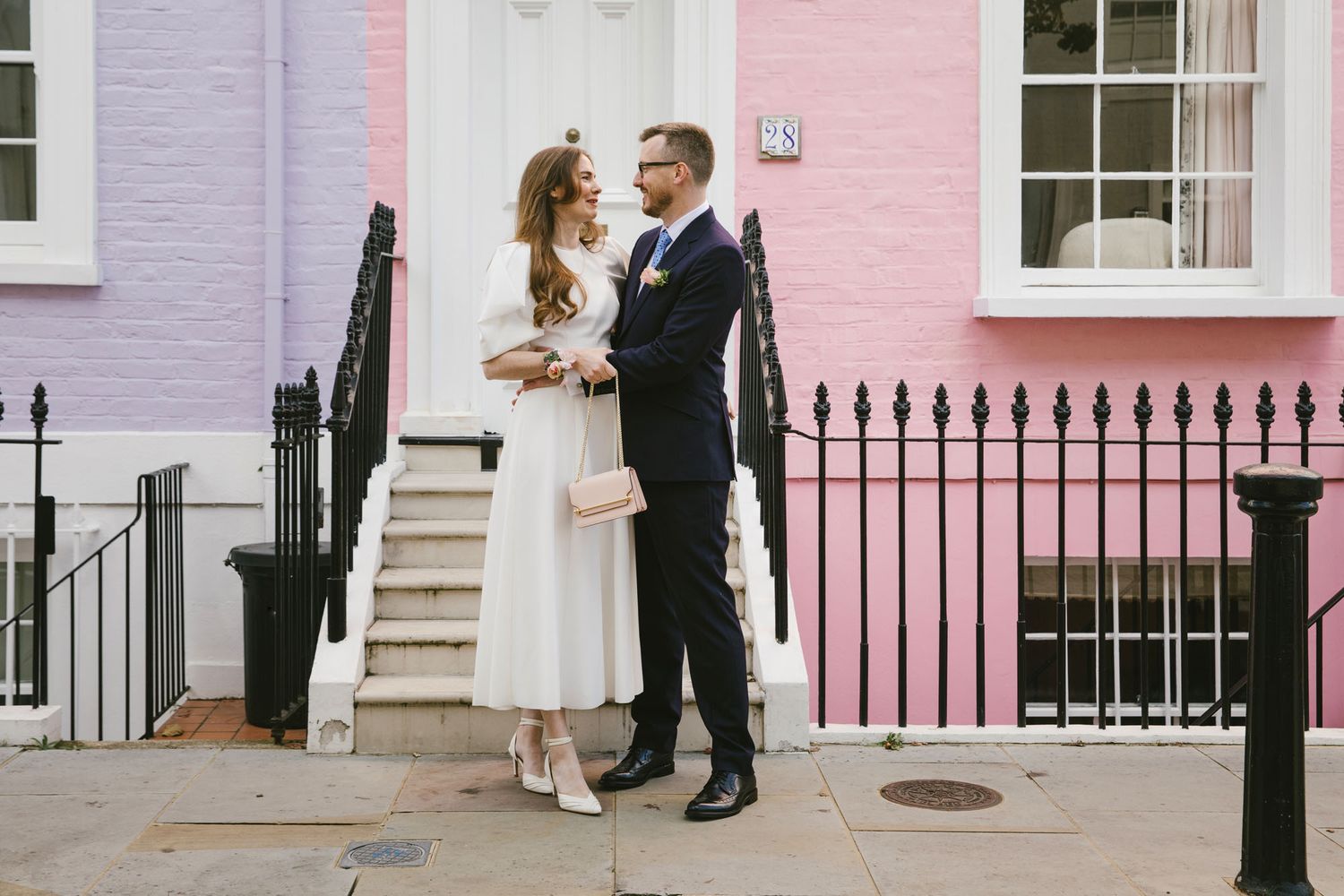 Couple in wedding attire standing on a colorful street with pink and purple buildings in London.