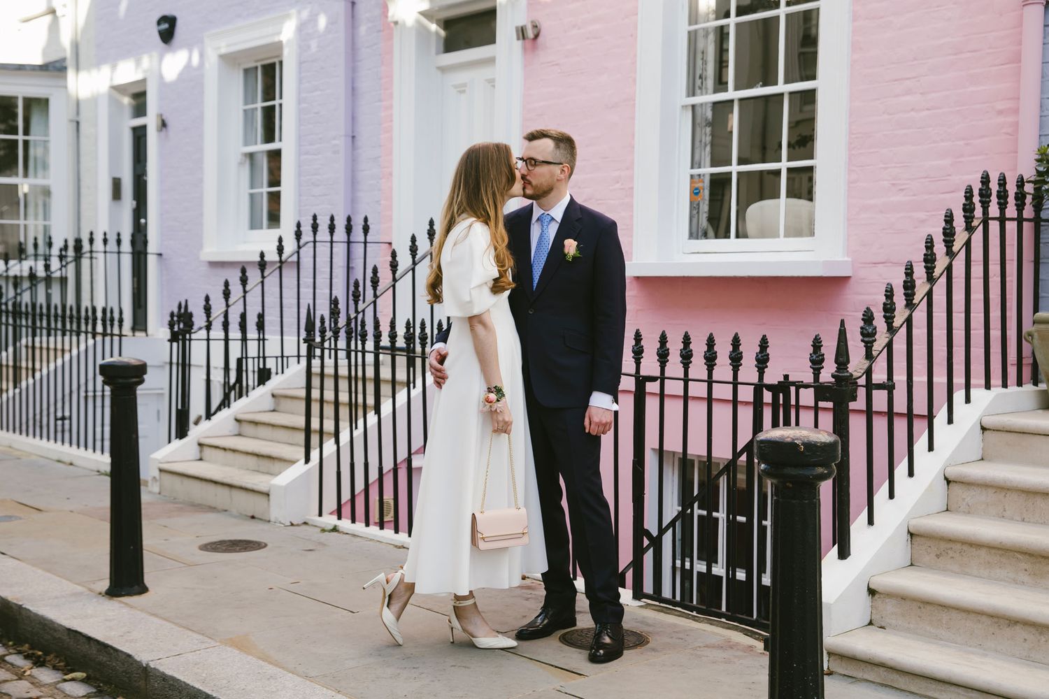 Couple in wedding attire sharing a romantic moment on a London street beside a pink building.
