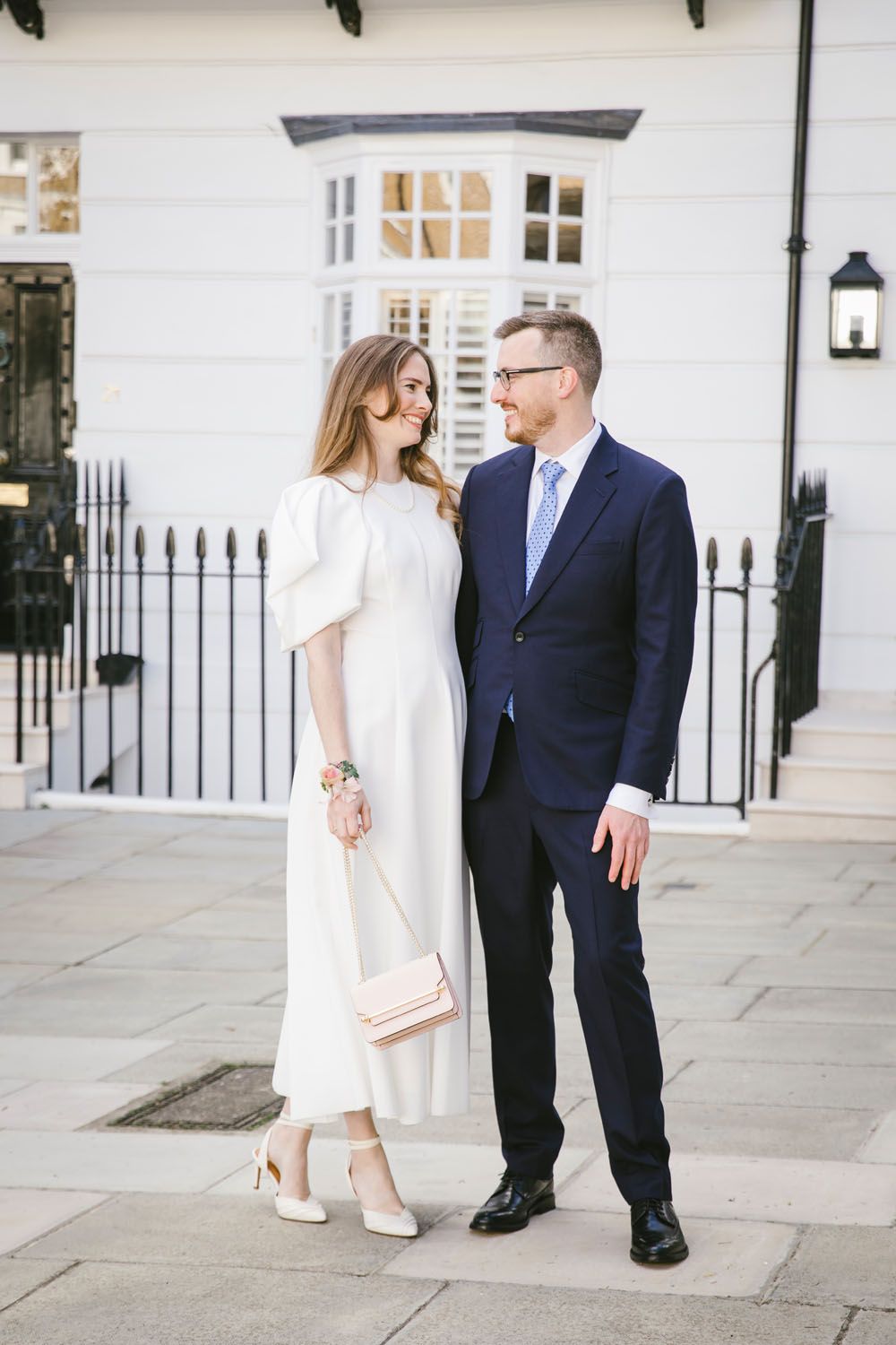 Elegant couple in wedding attire smiling at each other outside a white Georgian building.