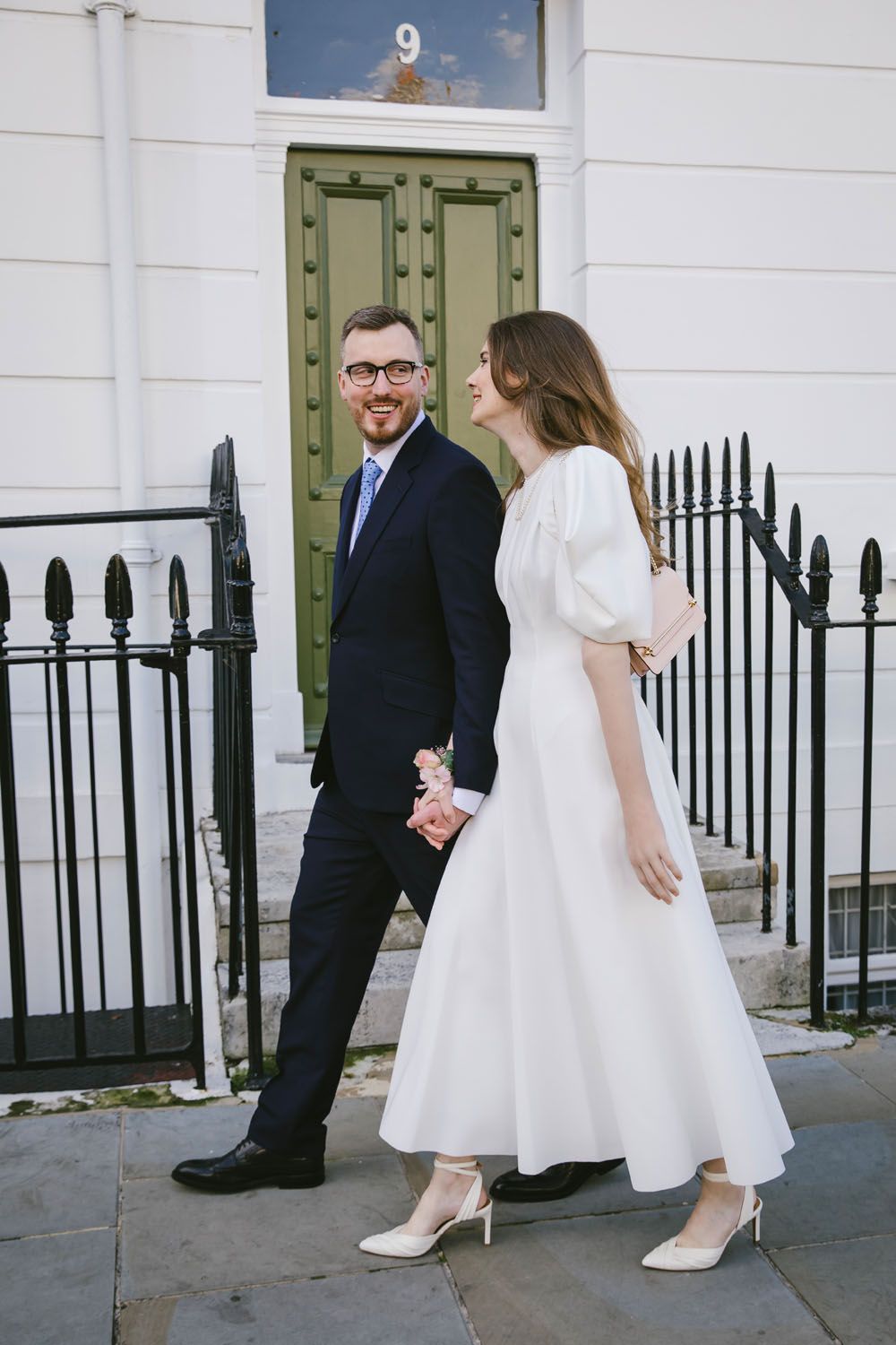 Smiling couple in wedding attire holding hands walking past a green door on a London street.