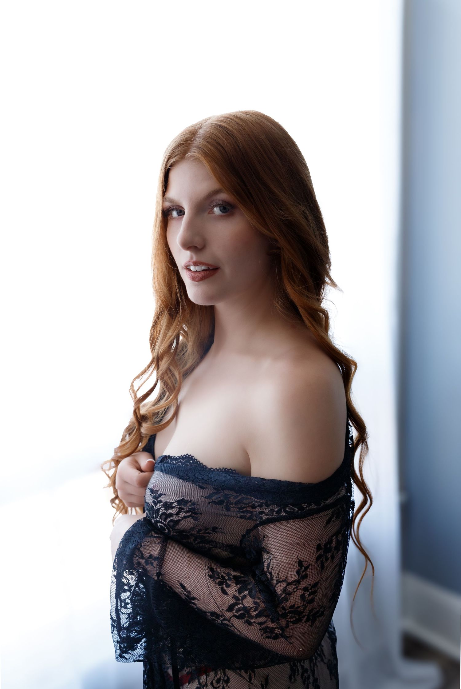 Tattooed bride in white lace lingerie and veil poses by a window in soft natural light.