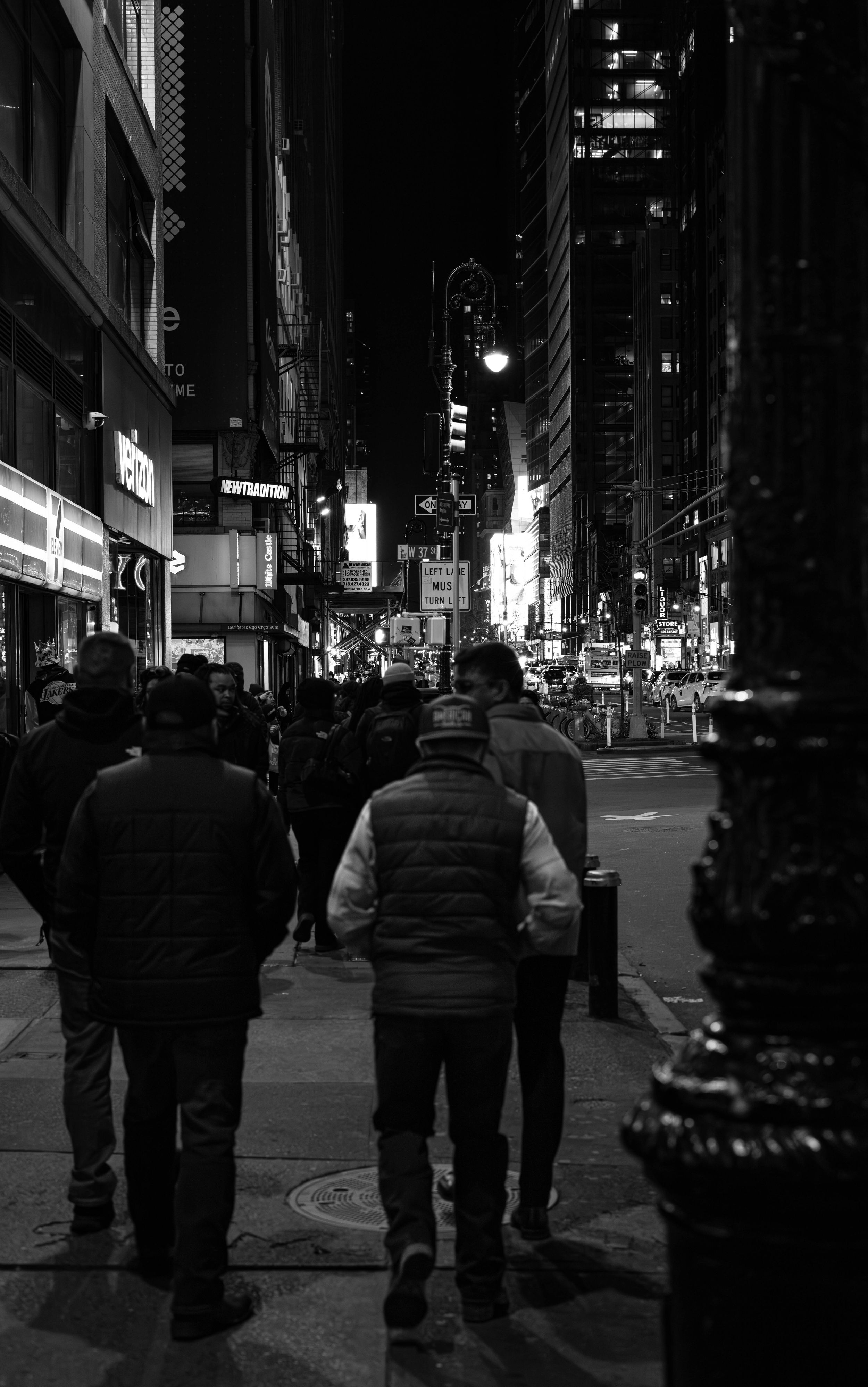 Black and white photo of people walking on a busy New York City sidewalk at night with illuminated buildings.