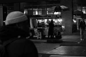 Black and white night street photo of a musician performing near a lit food cart while a person in a beanie watches.