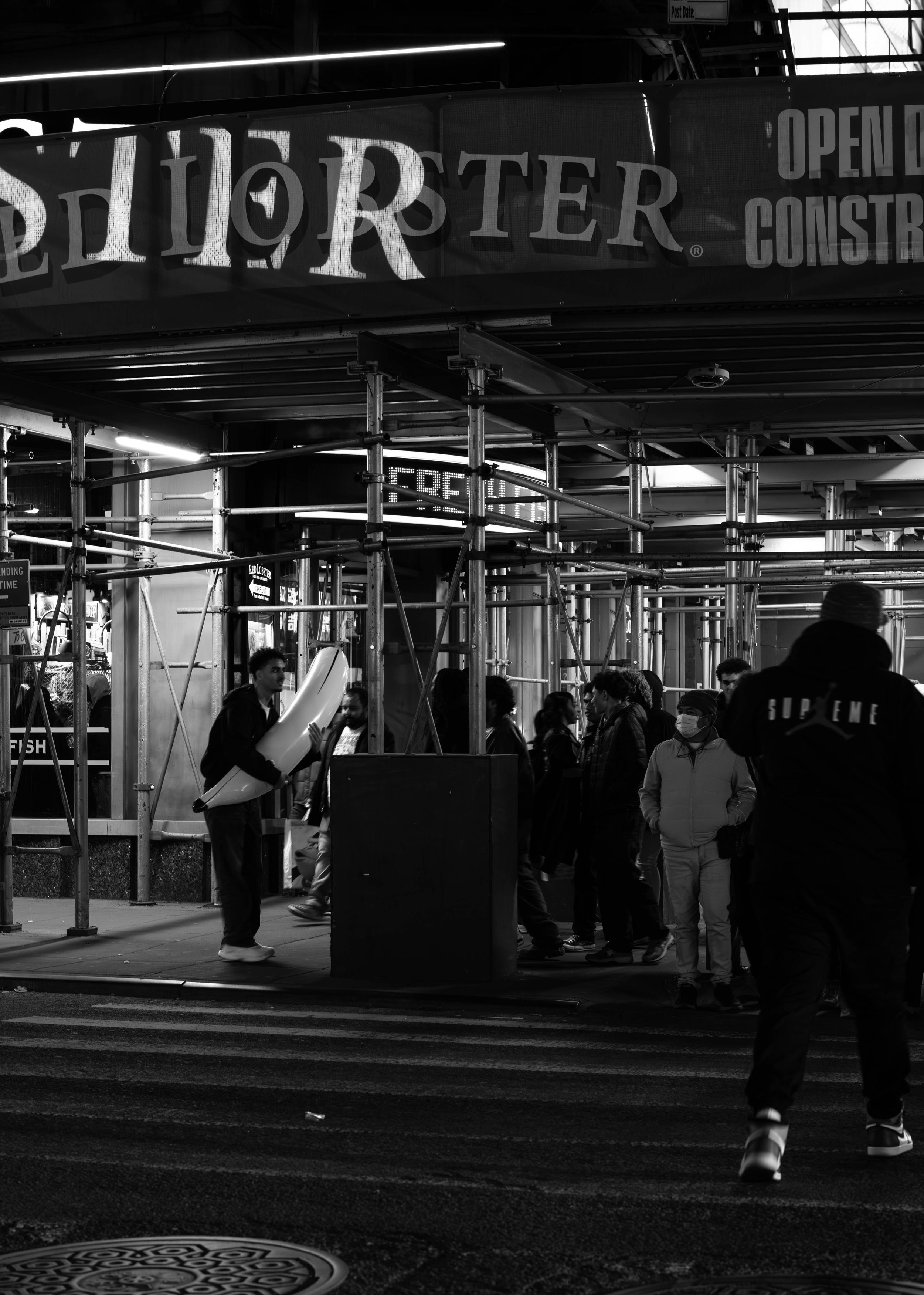Black and white street photo of people gathered outside a construction site at night near a Supreme store.